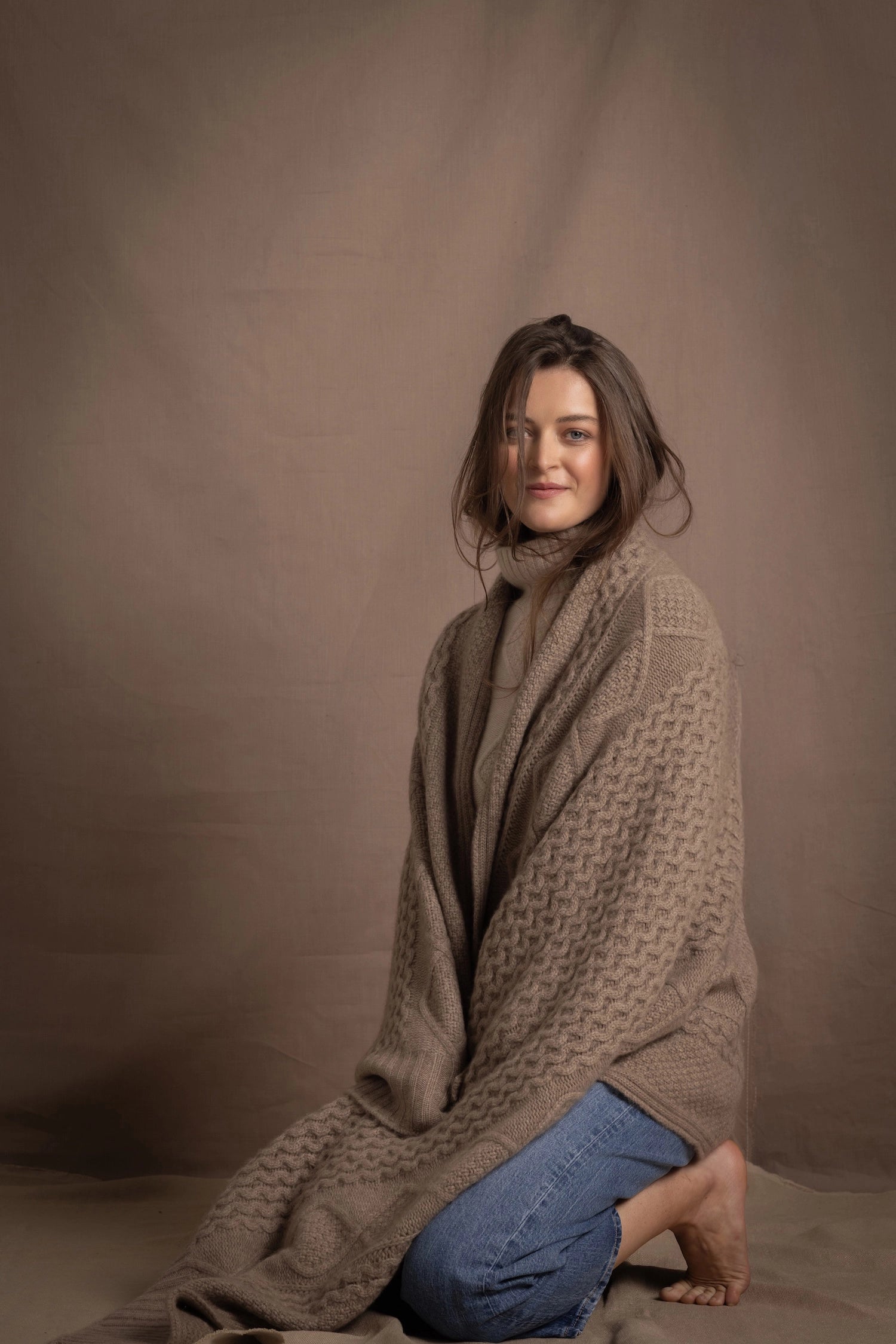 A lady wears a pure cashmere aran blanket in taupe over a beige aran sweater