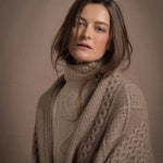 A lady wears a pure cashmere aran blanket in taupe over a beige aran sweater