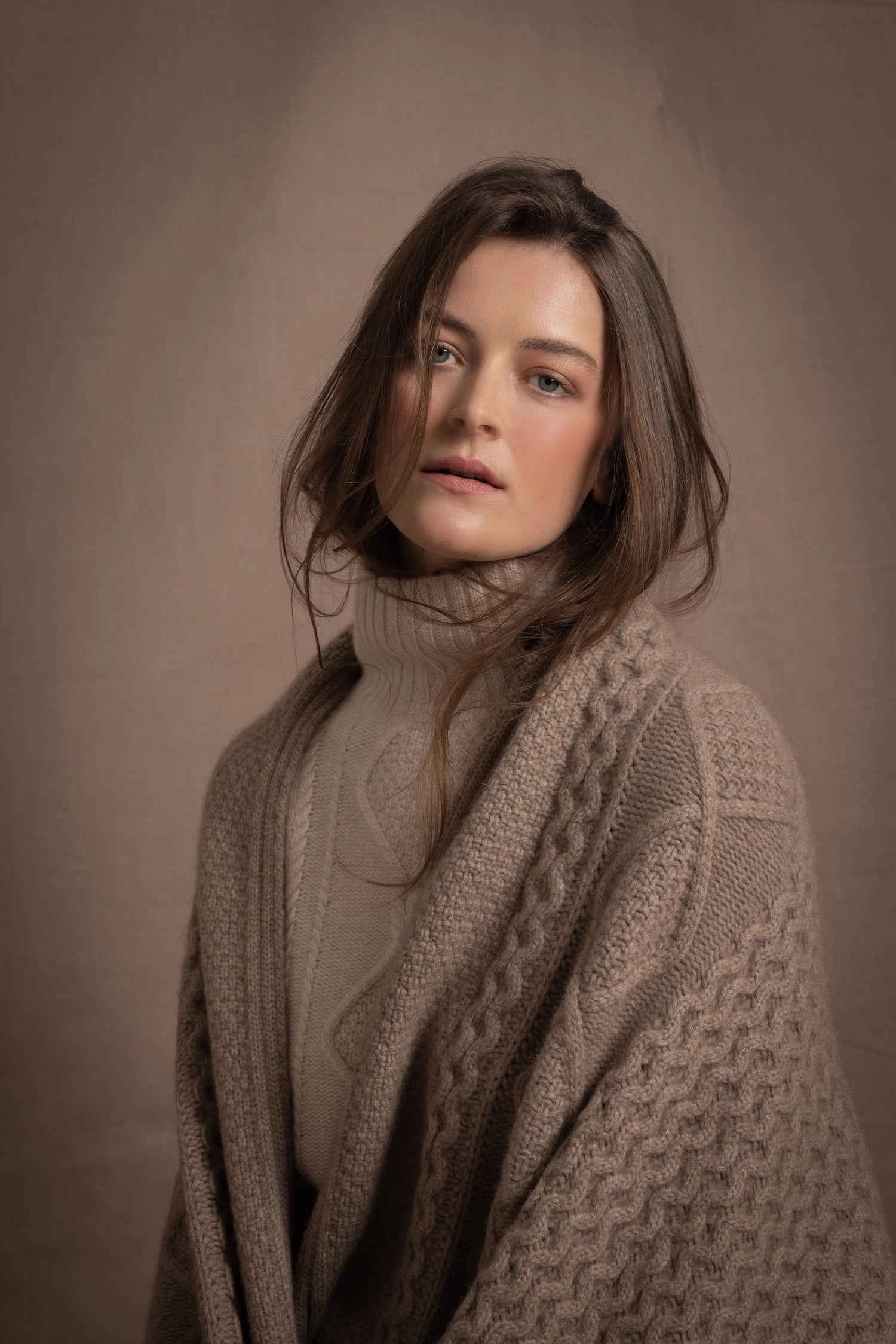 A lady wears a pure cashmere aran blanket in taupe over a beige aran sweater