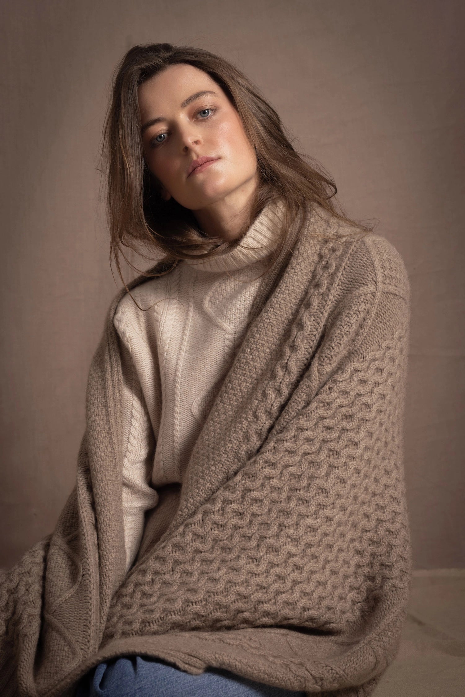 A lady wears a pure cashmere aran blanket in taupe over a beige aran sweater