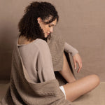 A lady wears a pure cashmere aran blanket in taupe over a beige cashmere sweater