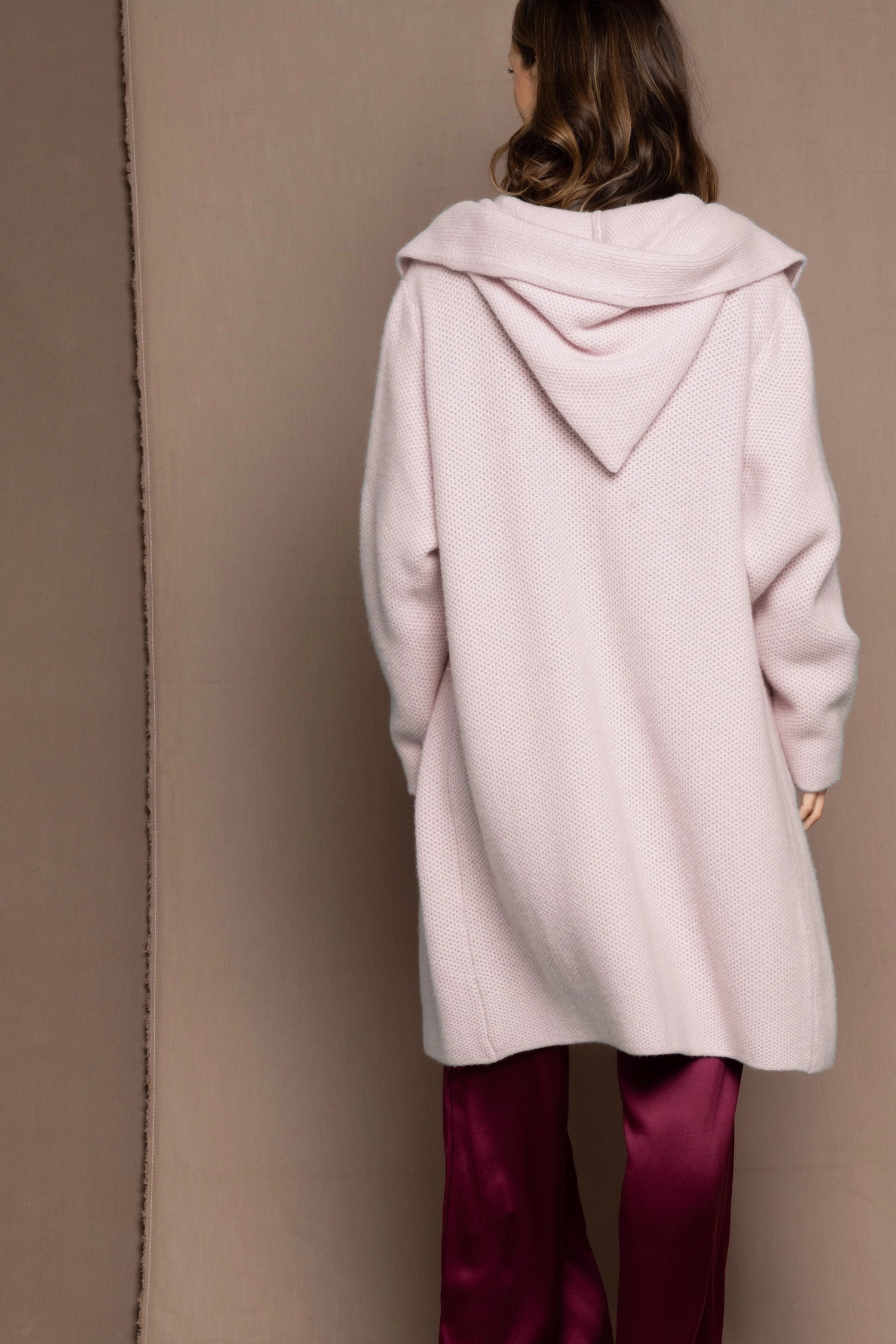 woman wearing a long cashmere coat cardigan in pale pink over burgundy silk trousers
