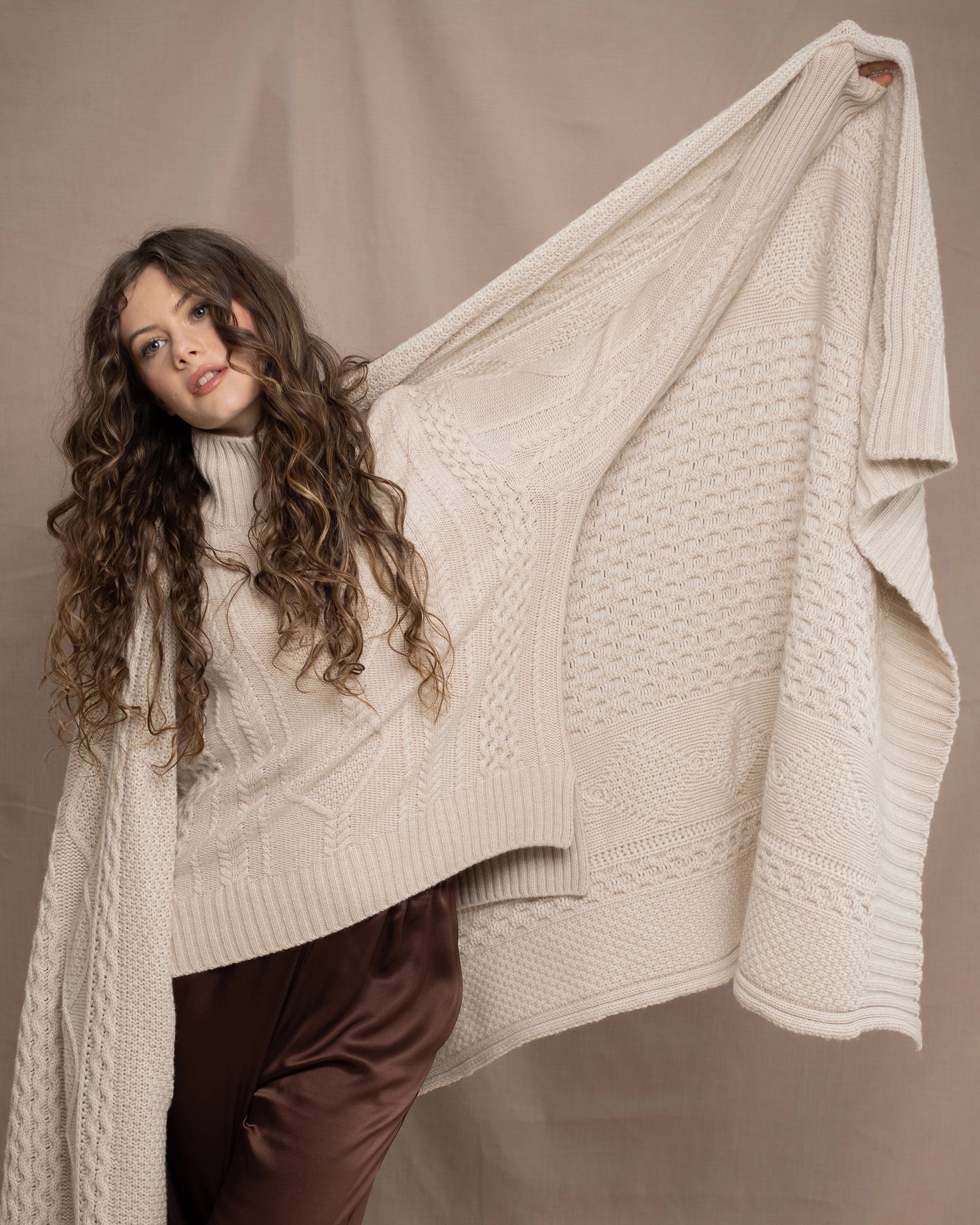 A woman wears a traditional handknit cashmere Aran Sweater Jumper from Ireland in Cream holding a matching cashmere aran throw blanket in cream with a chocolate brown silk skirt