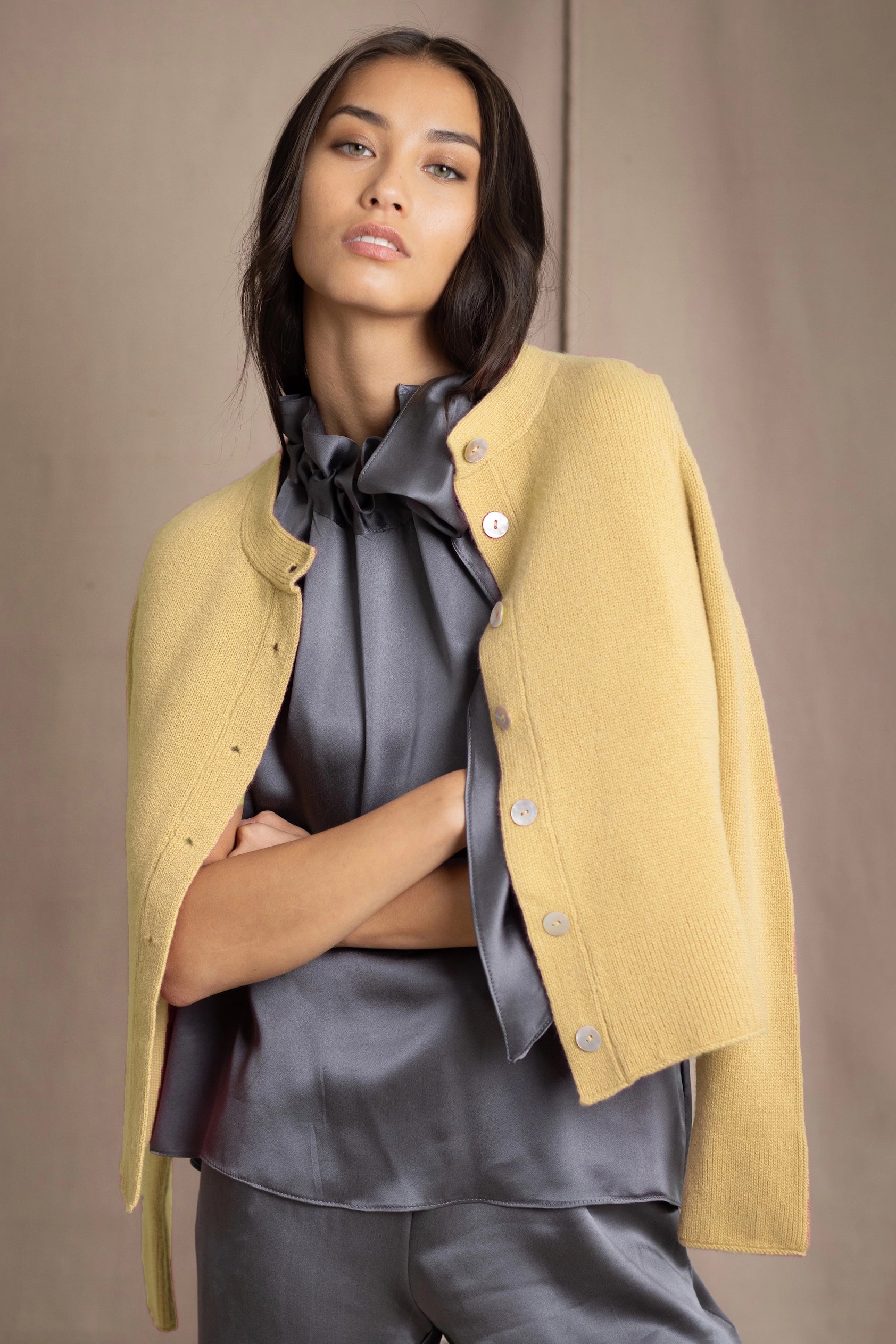 A woman wears a pale yellow cropped button-up cashmere cardigan over a dark grey silk blouse and matching grey silk trousers