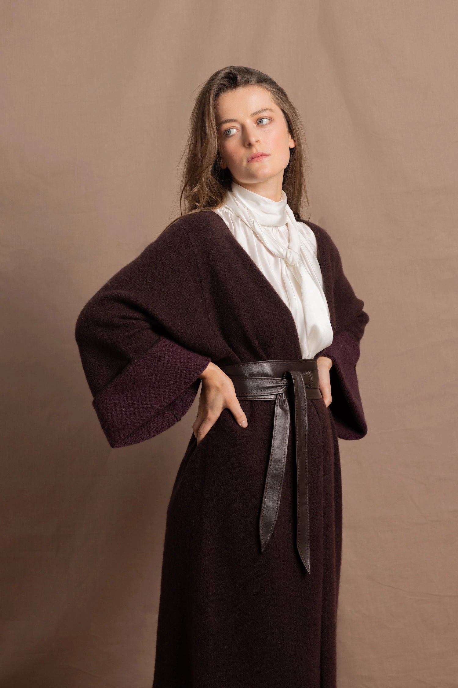 A woman wears a 100% silk pussybow sleeveless blouse in white with a chocolate brown and purple cashmere coat on top and cinched with leather obi belt