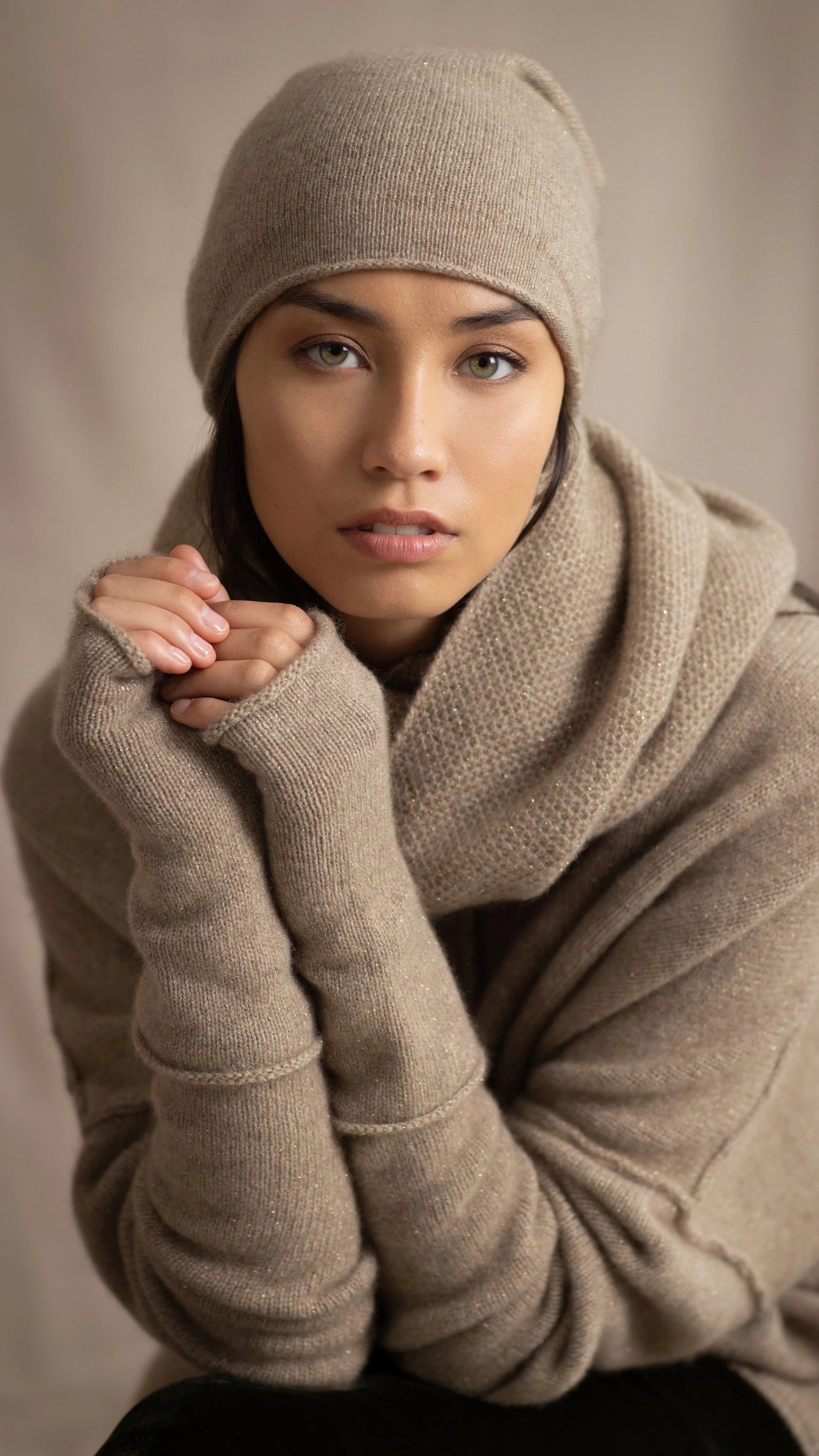 a woman wears a cashmere beanie hat, cashmere fingerless gloves, cashmere snood scarf and cashmere sweater in beige with gold sparkle