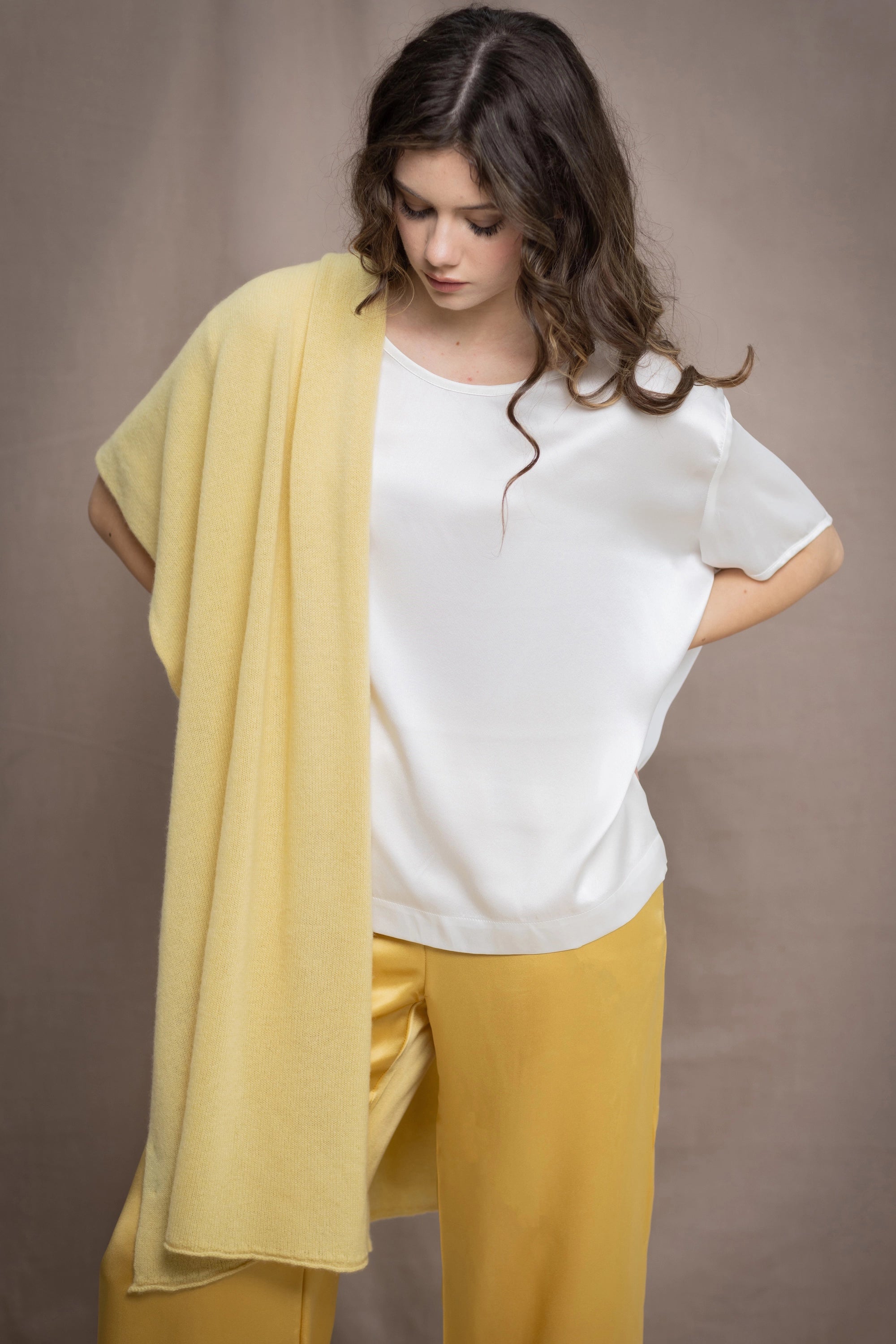 A woman wears a pale yellow cashmere travel wrap shawl over a silk tee in white and silk trousers in yellow