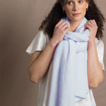 a woman wears a pale blue cashmere wrap scarf tied around her neck over a silk white tee and jeans