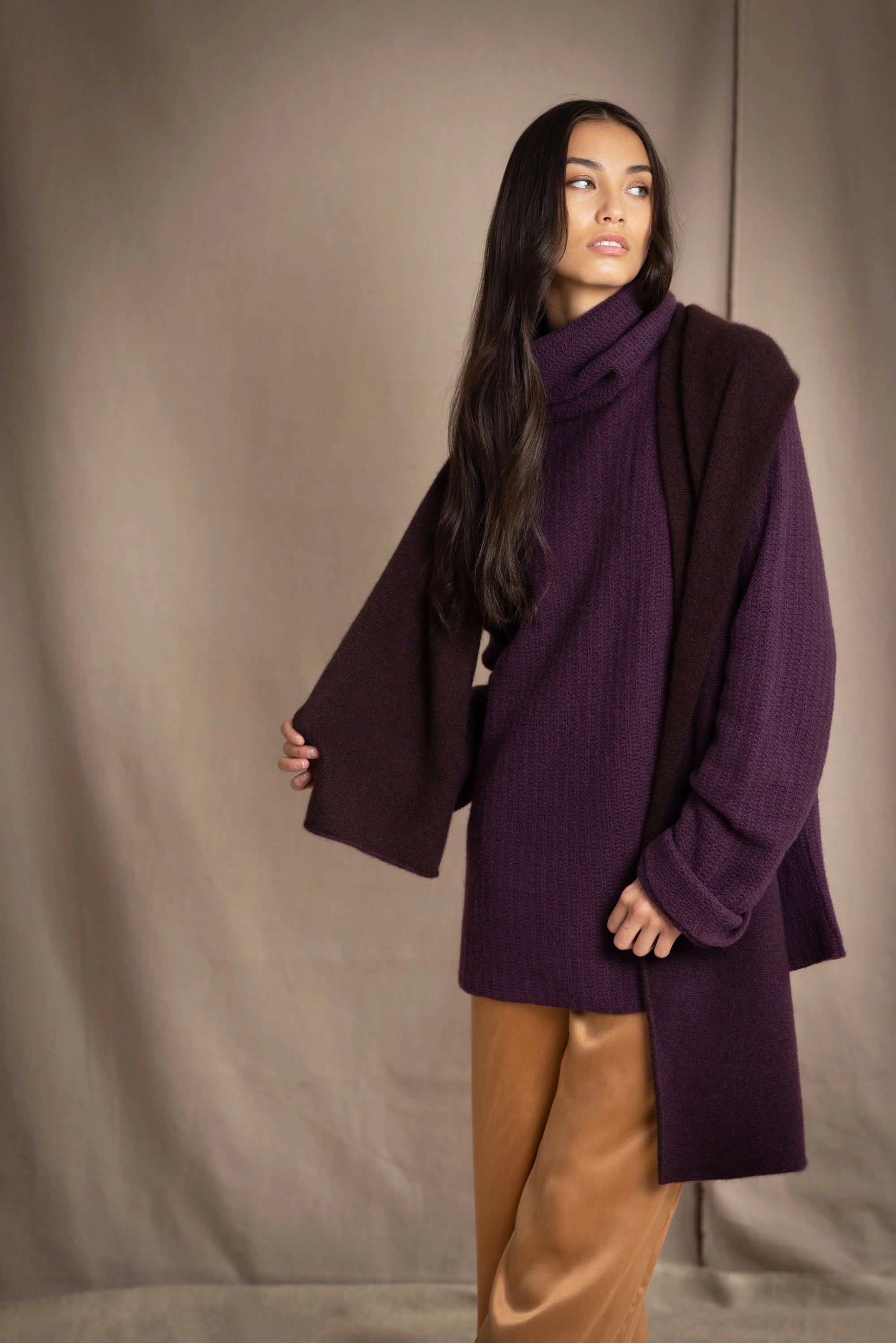 woman wears a reversible cashmere scarf in dark purple and dark brown on the other side worn over a dark purple cashmere sweater and bronze silk trousers