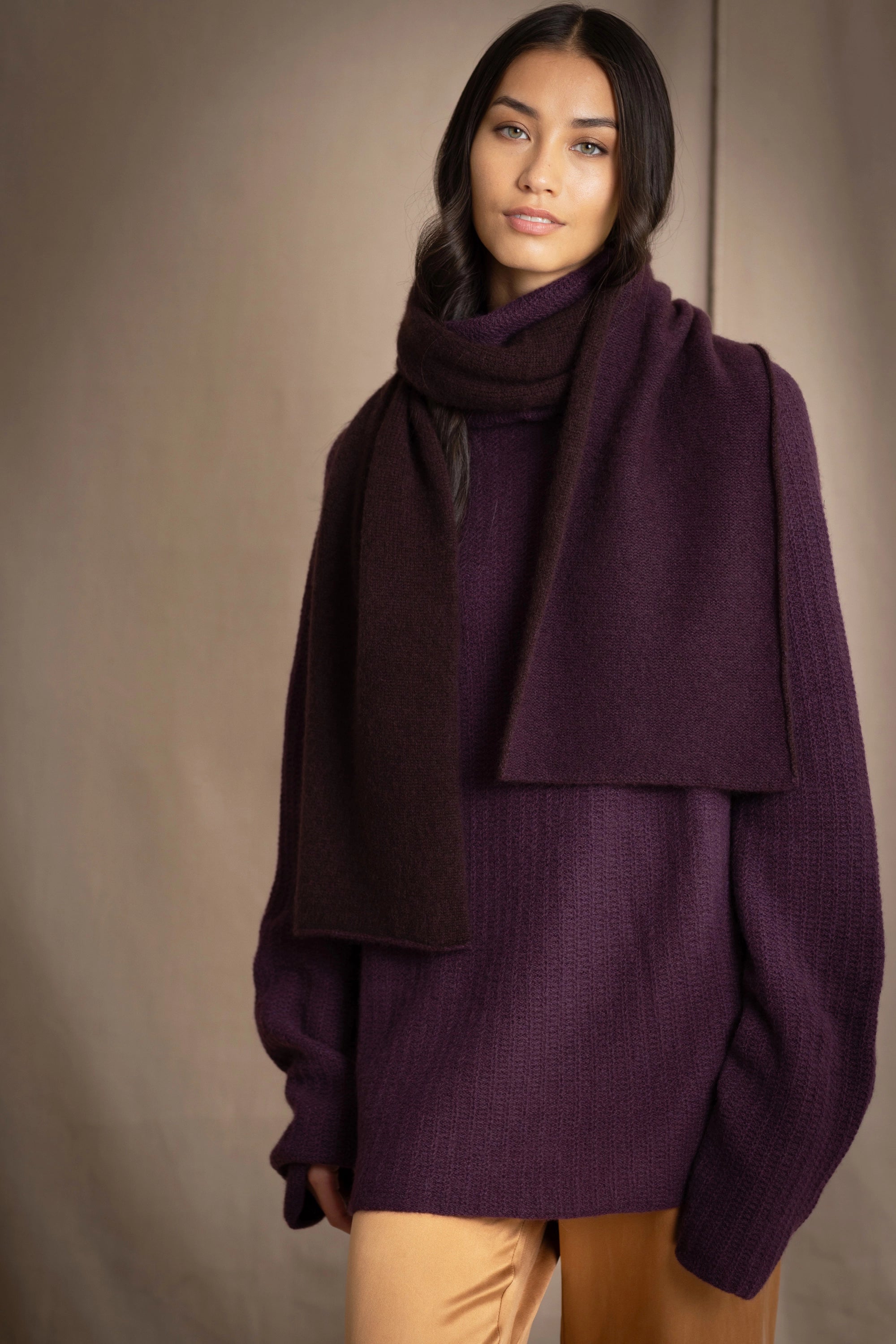 woman wears a reversible cashmere scarf in dark purple and dark brown on the other side worn over a dark purple cashmere sweater and bronze silk trousers