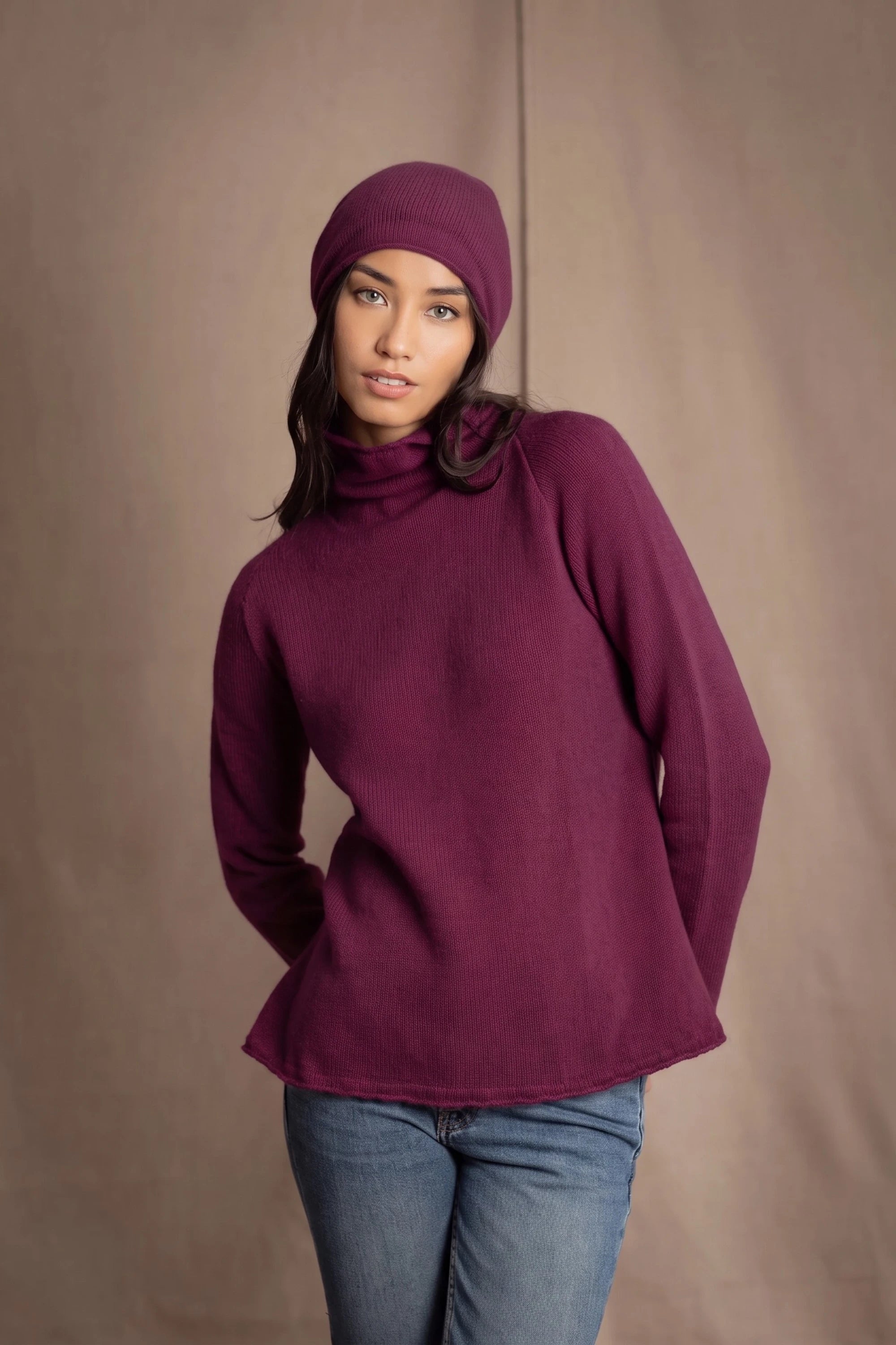 A woman wearing a burgundy cashmere turtleneck sweater, matching knit cashmere beanie, and blue jeans