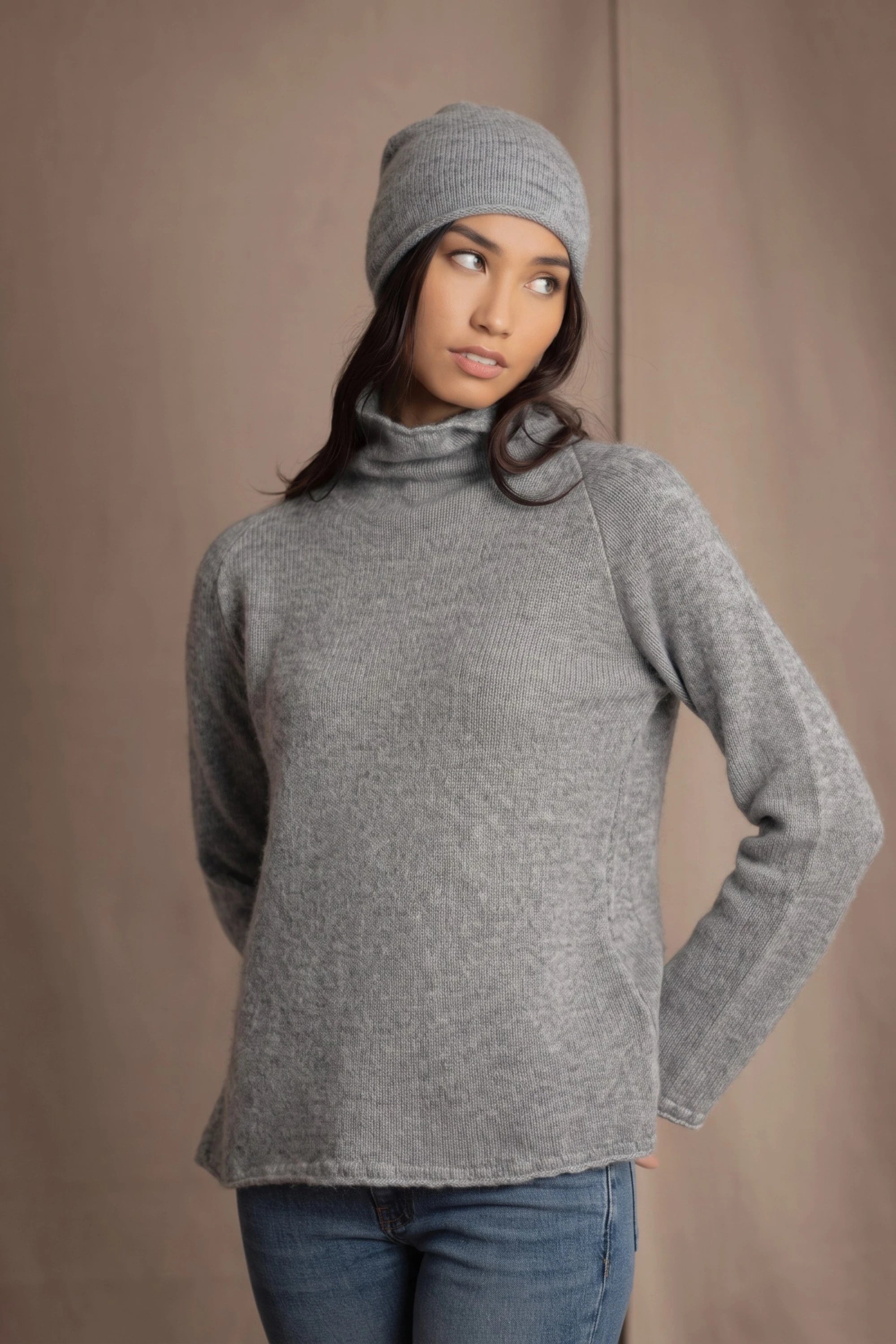 A woman wearing a light gray cashmere turtleneck sweater, matching knit cashmere beanie, and blue jeans