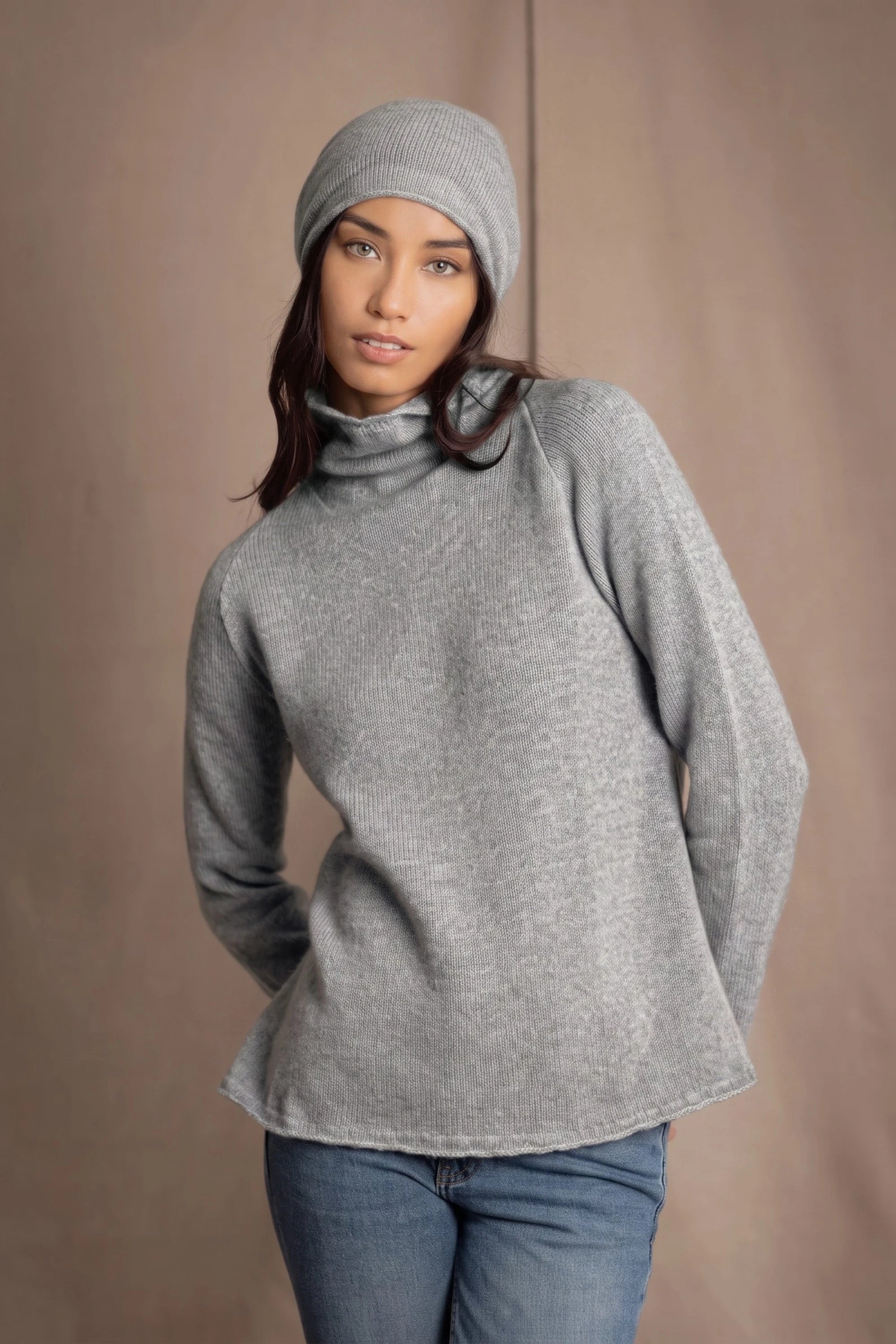 A woman wearing a light gray cashmere turtleneck sweater, matching knit cashmere beanie, and blue jeans