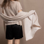 A woman wears a large cashmere shawl in beige over a matching cashmere tee and black silk shorts