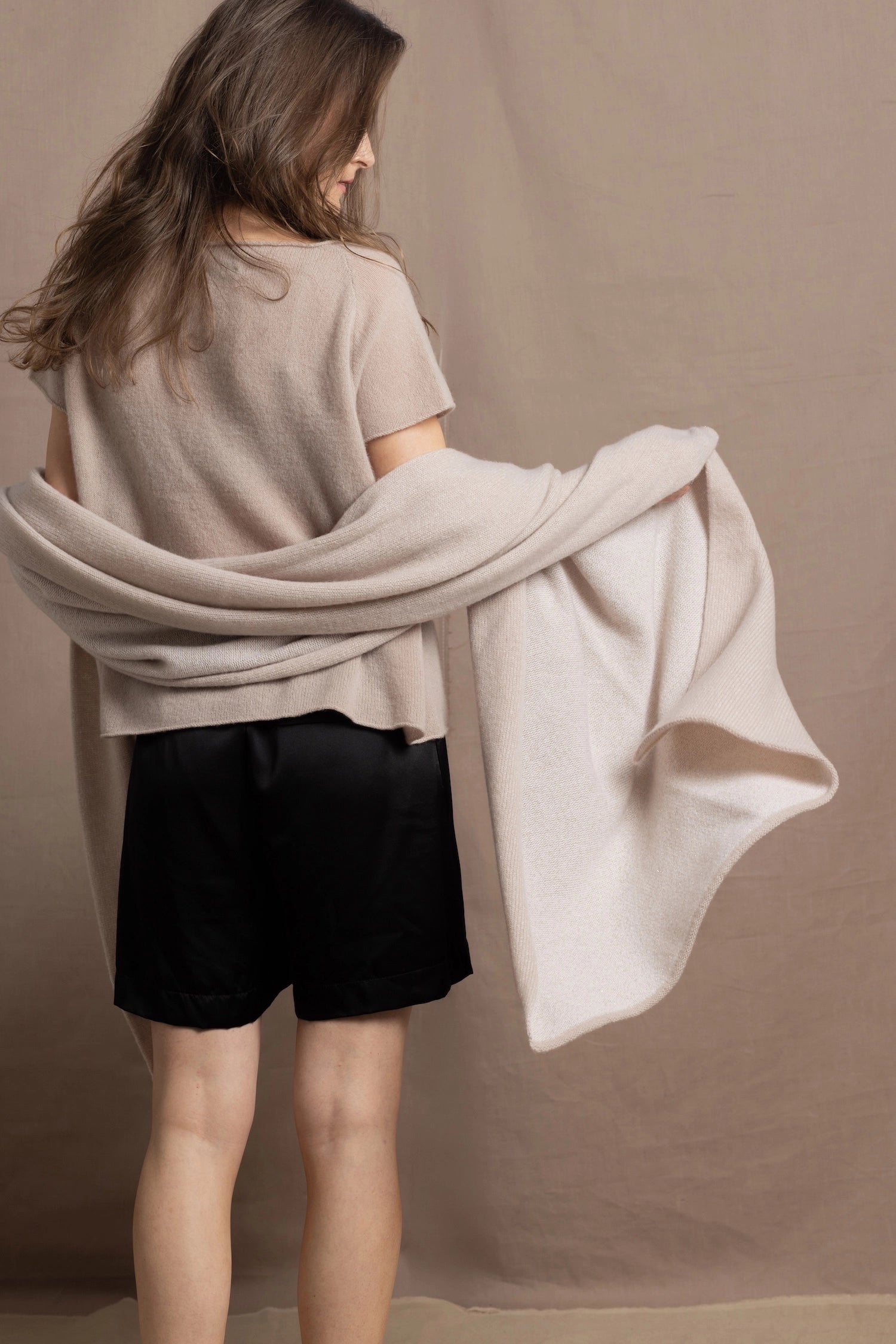A woman wears a large cashmere shawl in beige over a matching cashmere tee and black silk shorts