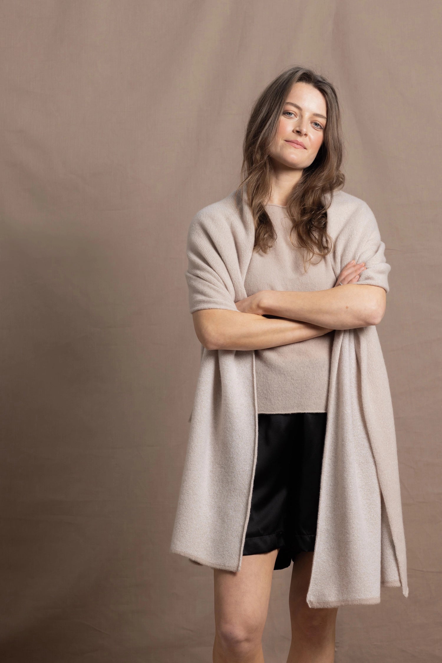 A woman wears a large cashmere shawl in beige over a matching cashmere tee and black silk shorts