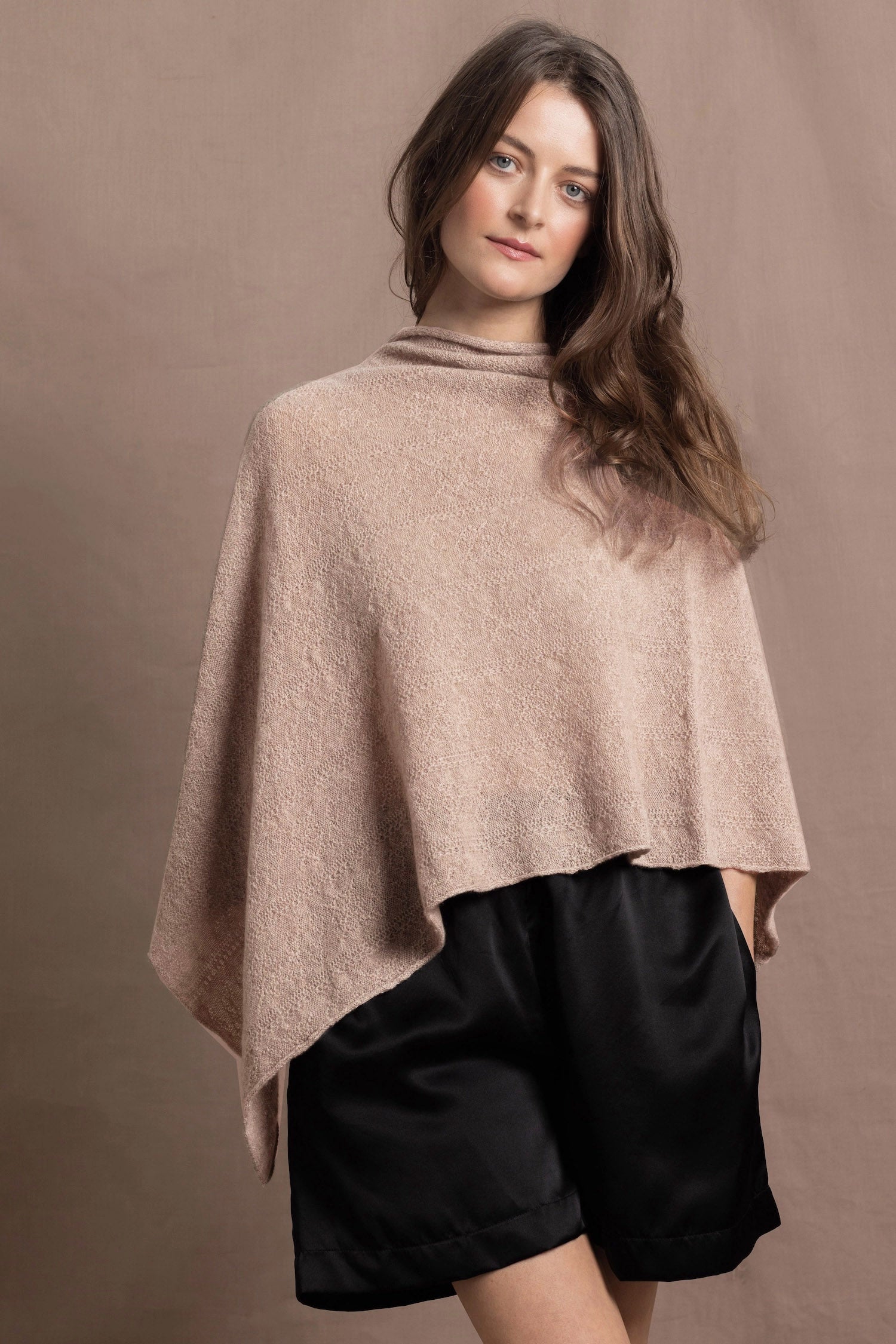 a woman wears a sand coloured cashmere cape or poncho over black silk shorts
