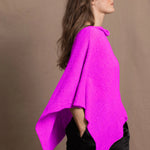 A woman wears a cashmere cape poncho in neon pink with black silk shorts