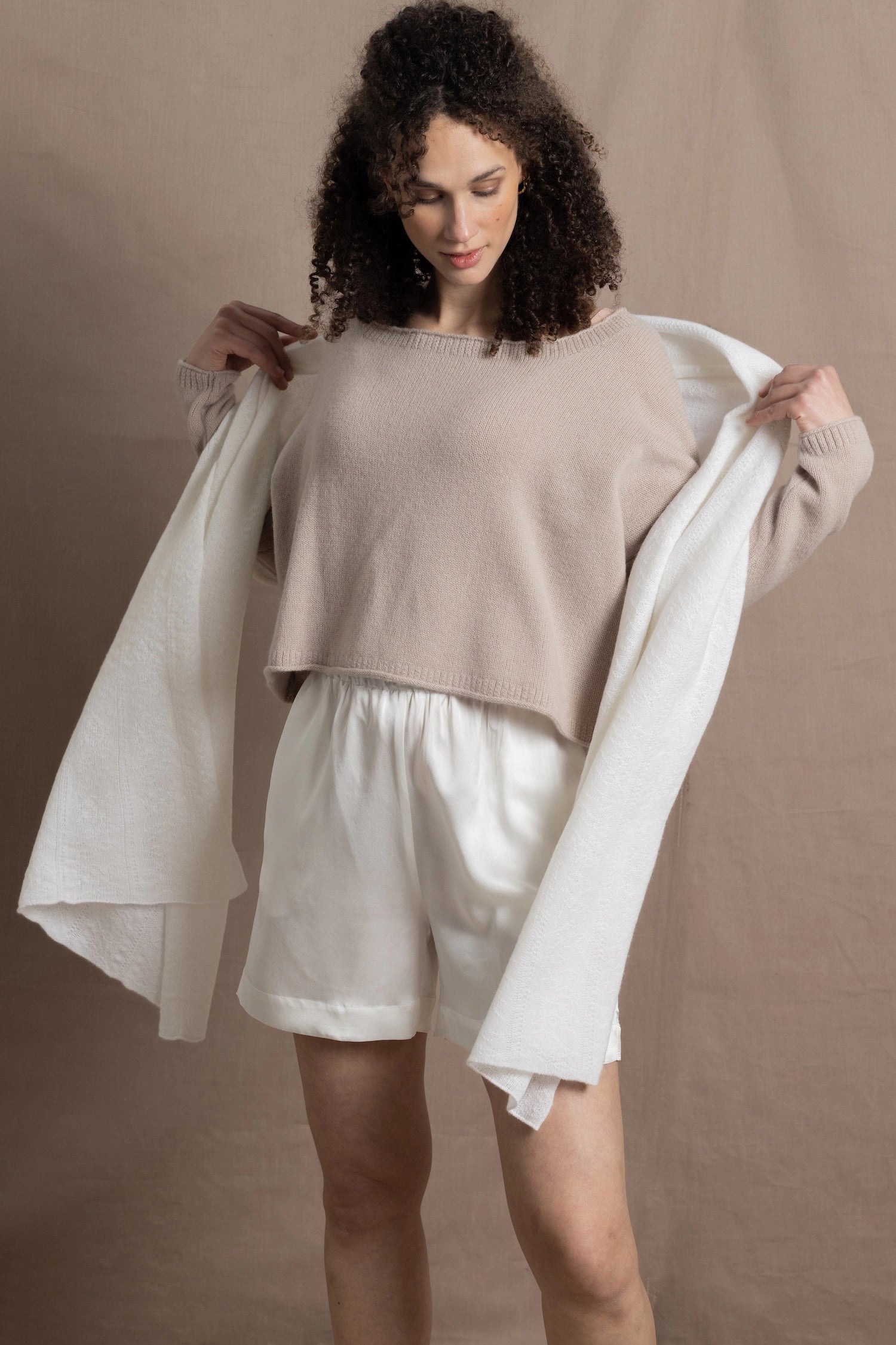 A woman wears a cropped cashmere sweater in beige with white silk shorts and matching cashmere wrap shawl
