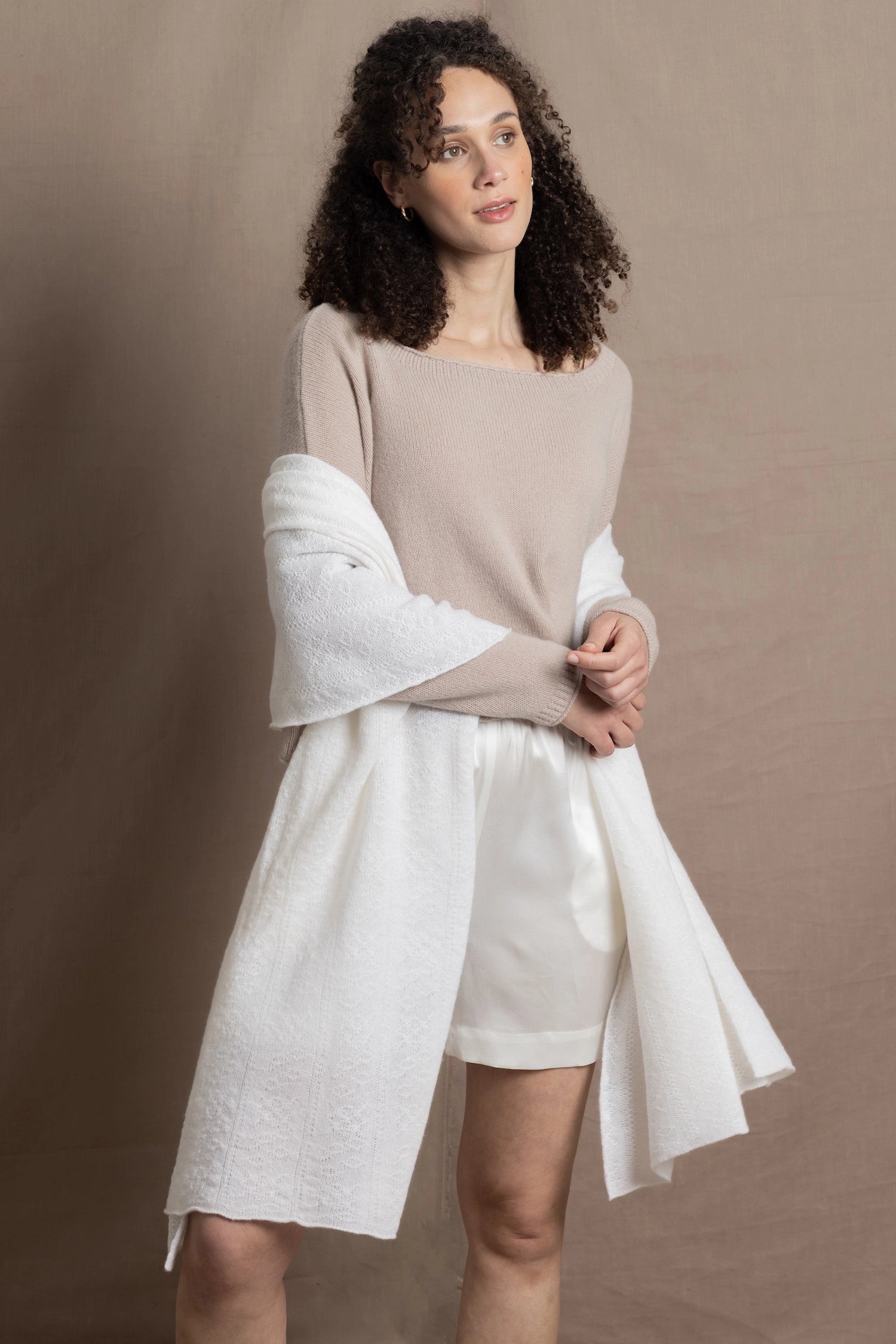 A lady wears a pure cashmere pointelle wrap in white over a beige cropped cashmere sweater and white silk shorts