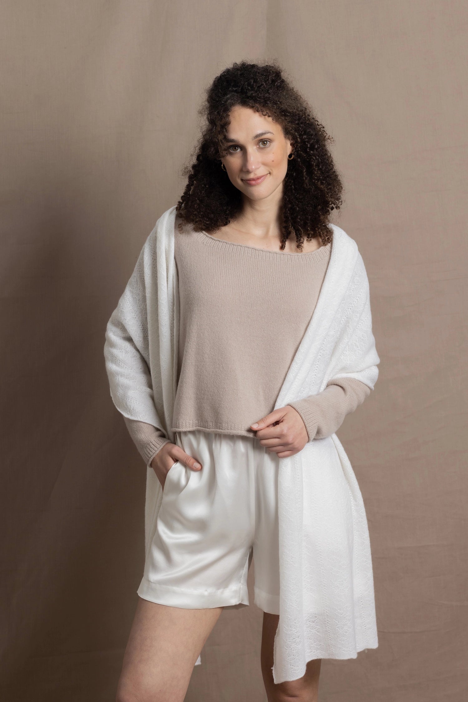 A lady wears a pure cashmere pointelle wrap in white over a beige cropped cashmere sweater and white silk shorts