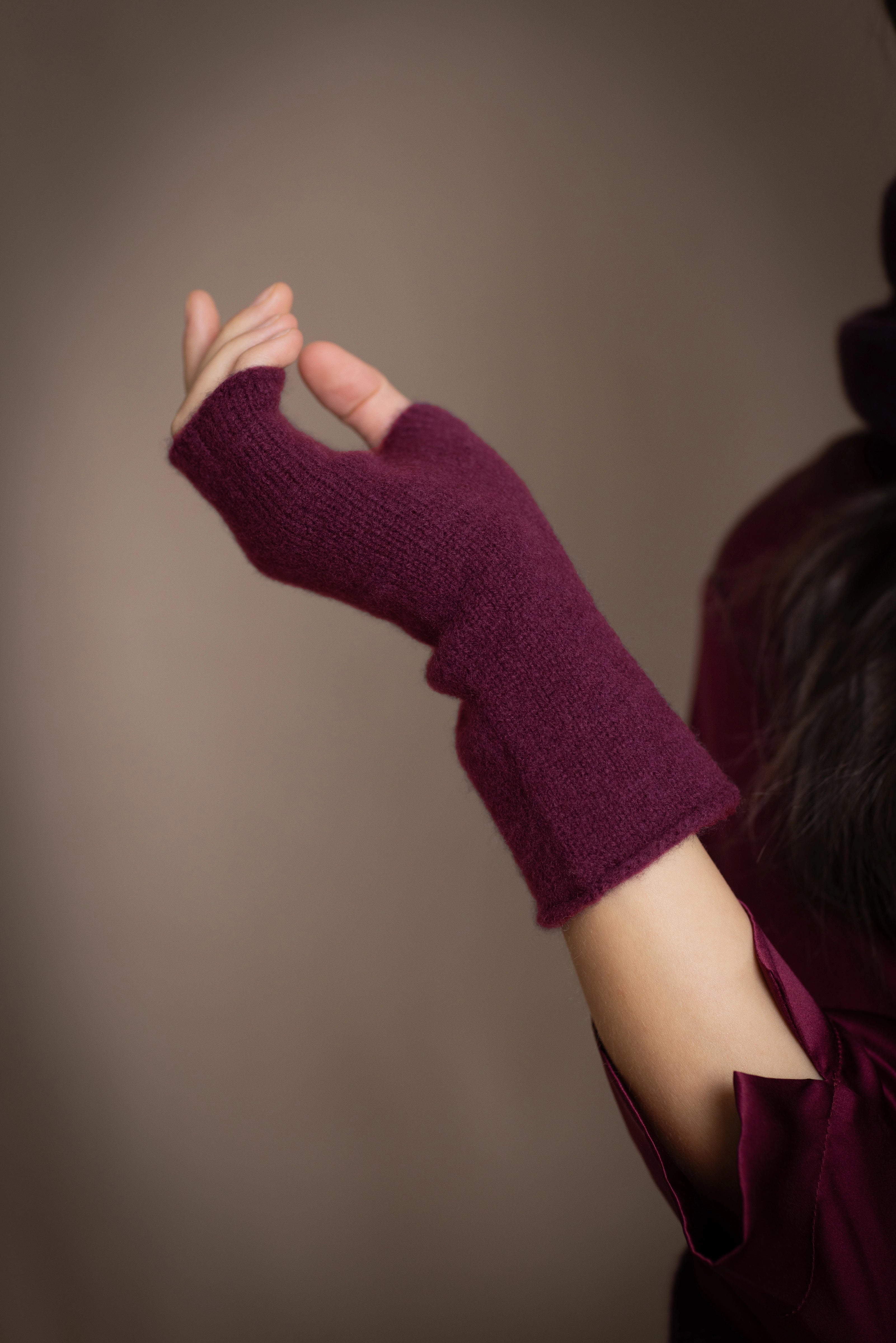 A close-up of a person's hand wearing fingerless cashmere gloves in deep plum burgundy