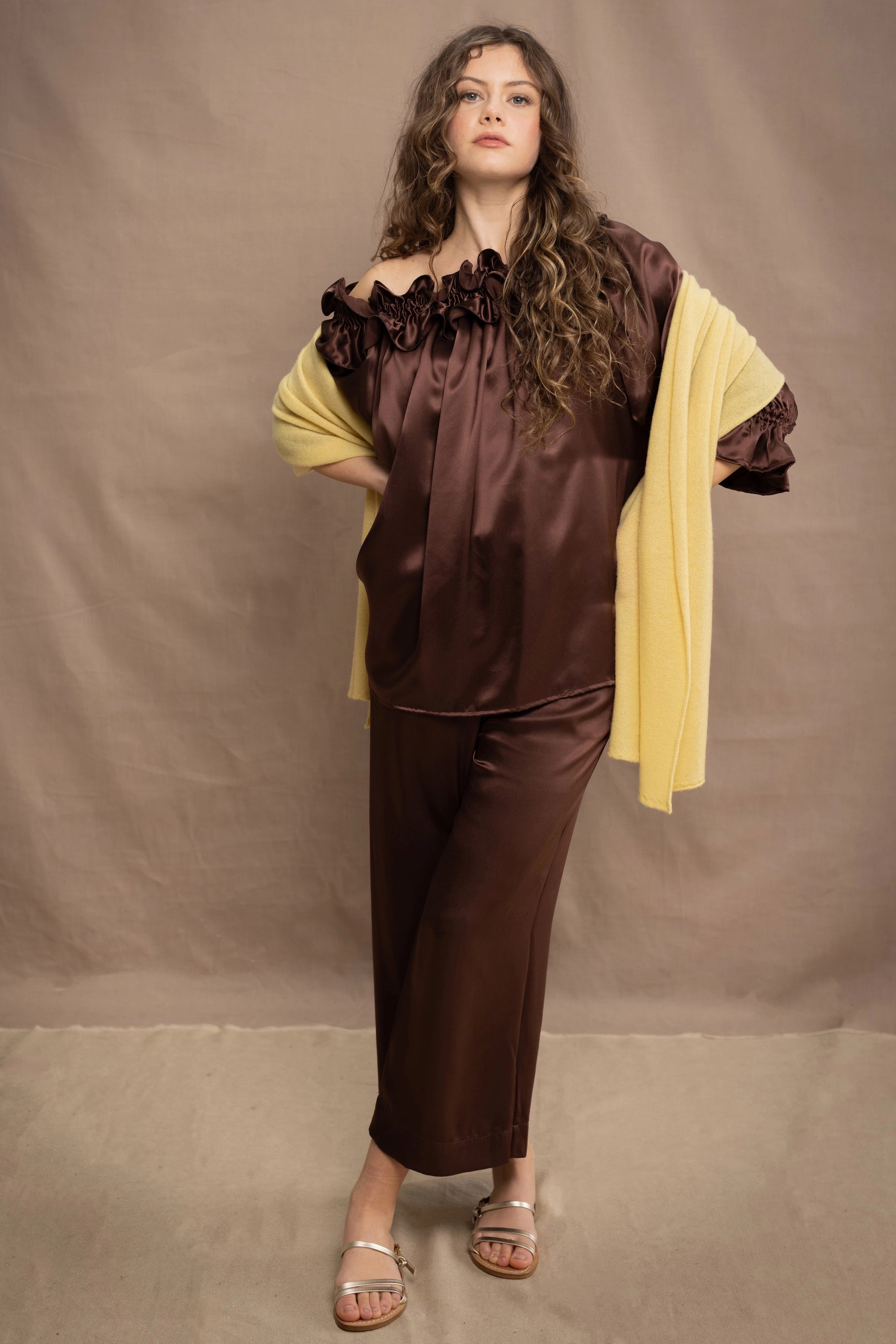 a woman wears a banana yellow cashmere wrap shawl over a ruffled silk blouse top in chocolate brown with matching trousers 