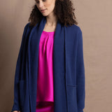 A woman wears an open style longer cashmere cardigan in blue worn over a bright pink 100% silk top and matching skirt