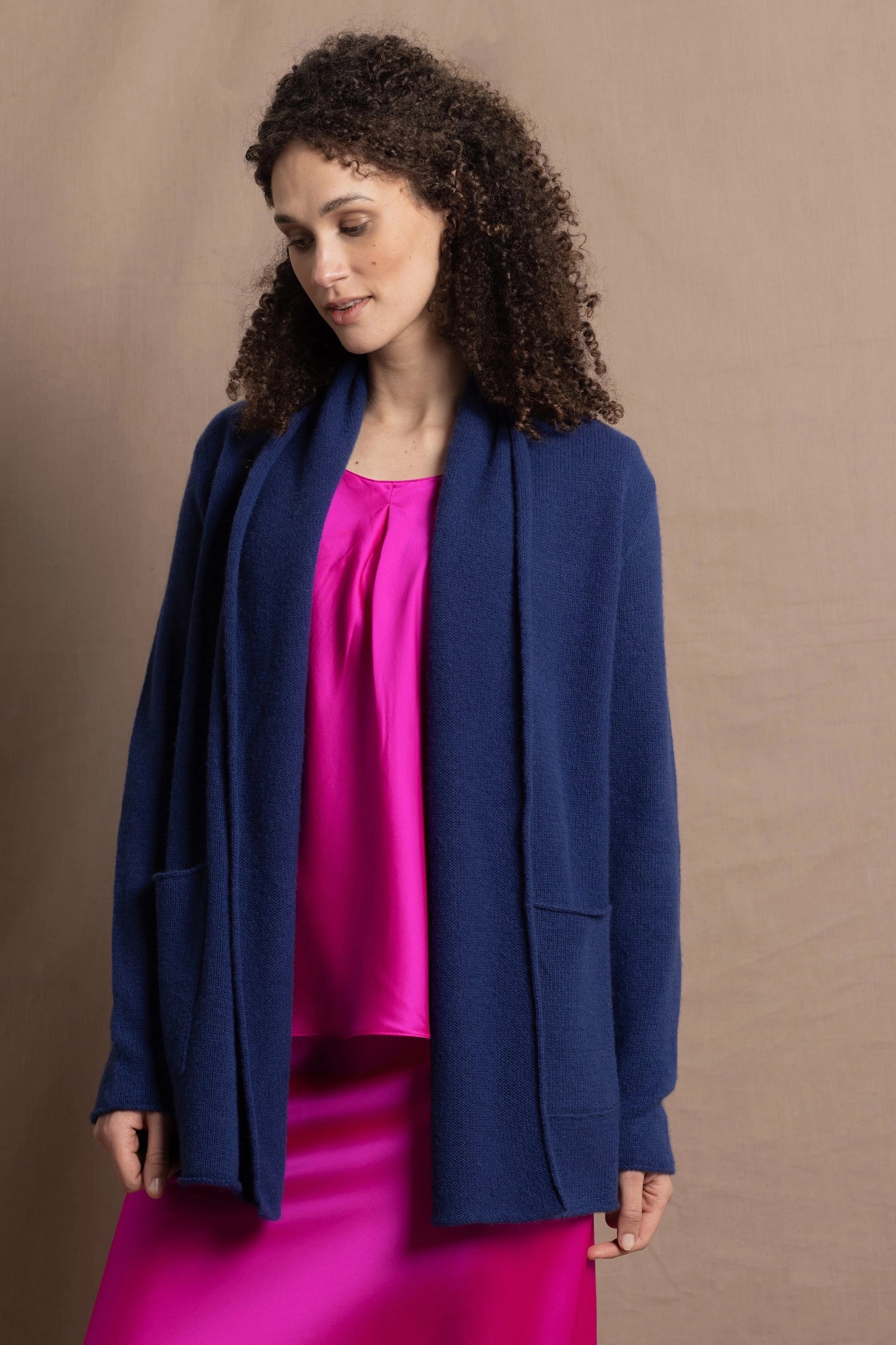 A woman wears an open style longer cashmere cardigan in blue worn over a bright pink 100% silk top and matching skirt
