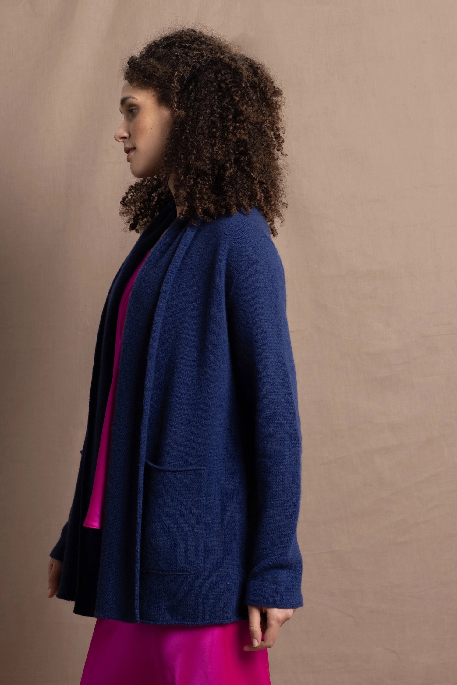 A woman wears an open style longer cashmere cardigan in blue worn over a bright pink 100% silk top and matching skirt