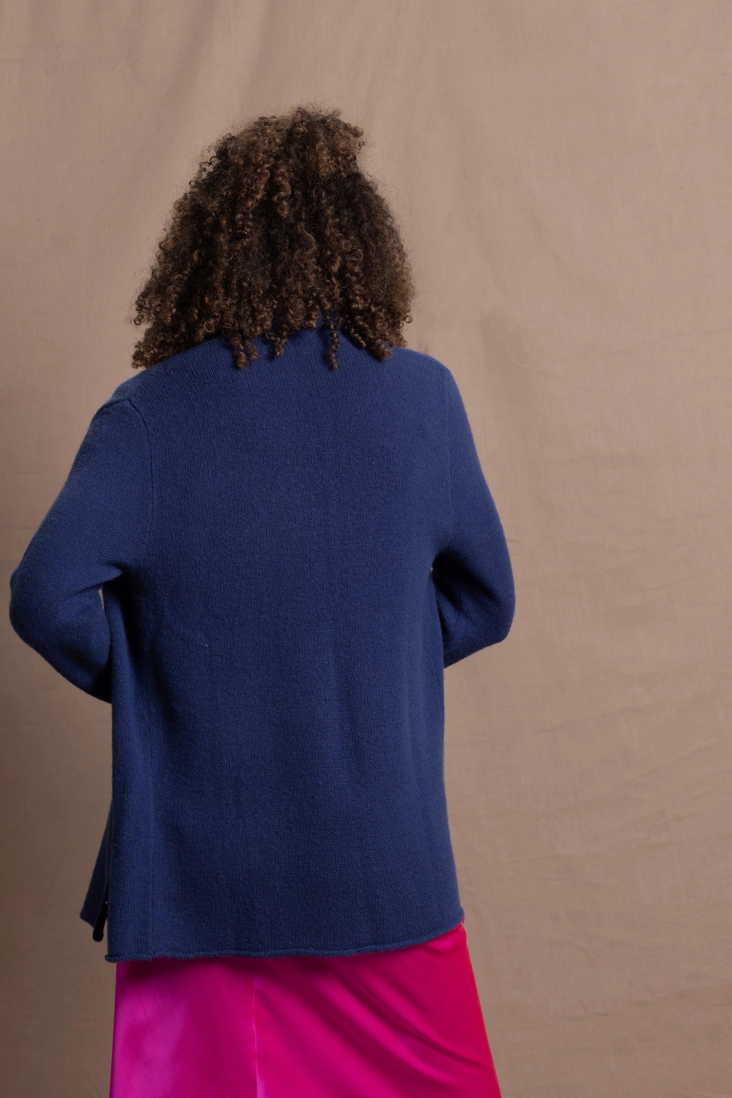 A woman wears an open style longer cashmere cardigan in blue worn over a bright pink 100% silk top and matching skirt