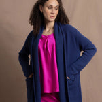 A woman wears an open style longer cashmere cardigan in blue worn over a bright pink 100% silk top and matching skirt