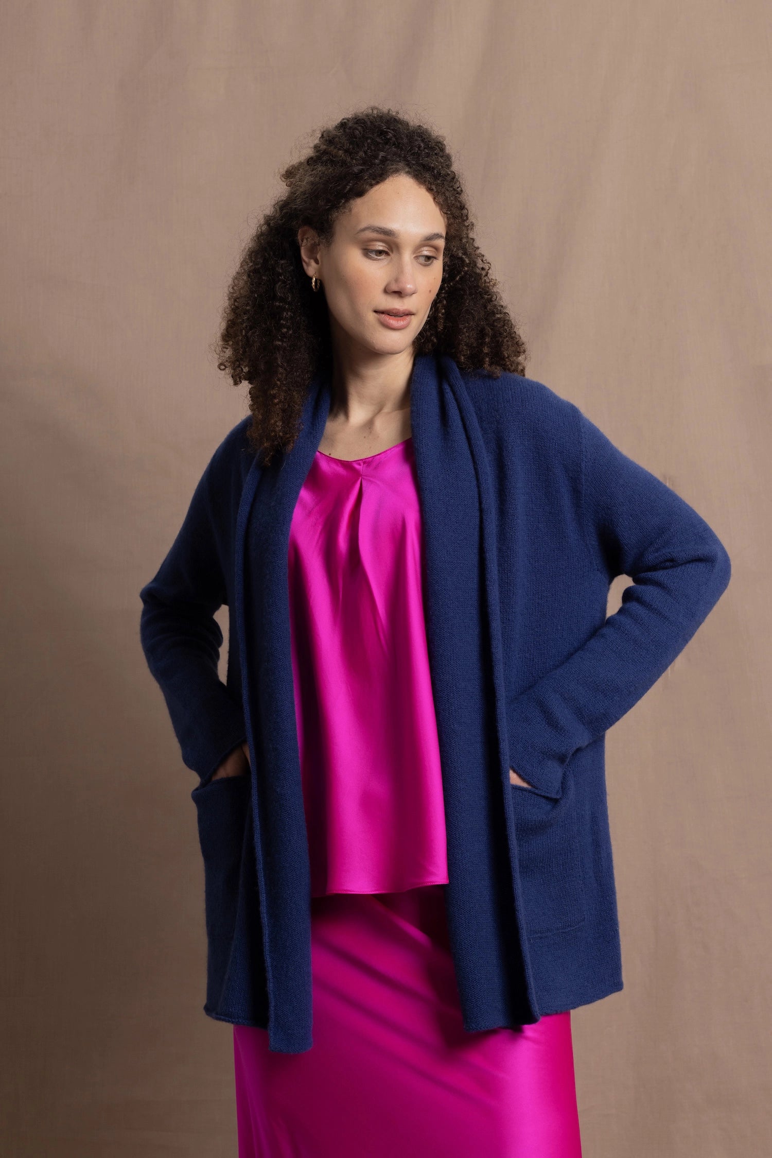 A woman wears an open style longer cashmere cardigan in blue worn over a bright pink 100% silk top and matching skirt