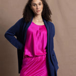 A woman wears an open style longer cashmere cardigan in blue worn over a bright pink 100% silk top and matching skirt