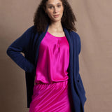 A woman wears an open style longer cashmere cardigan in blue worn over a bright pink 100% silk top and matching skirt