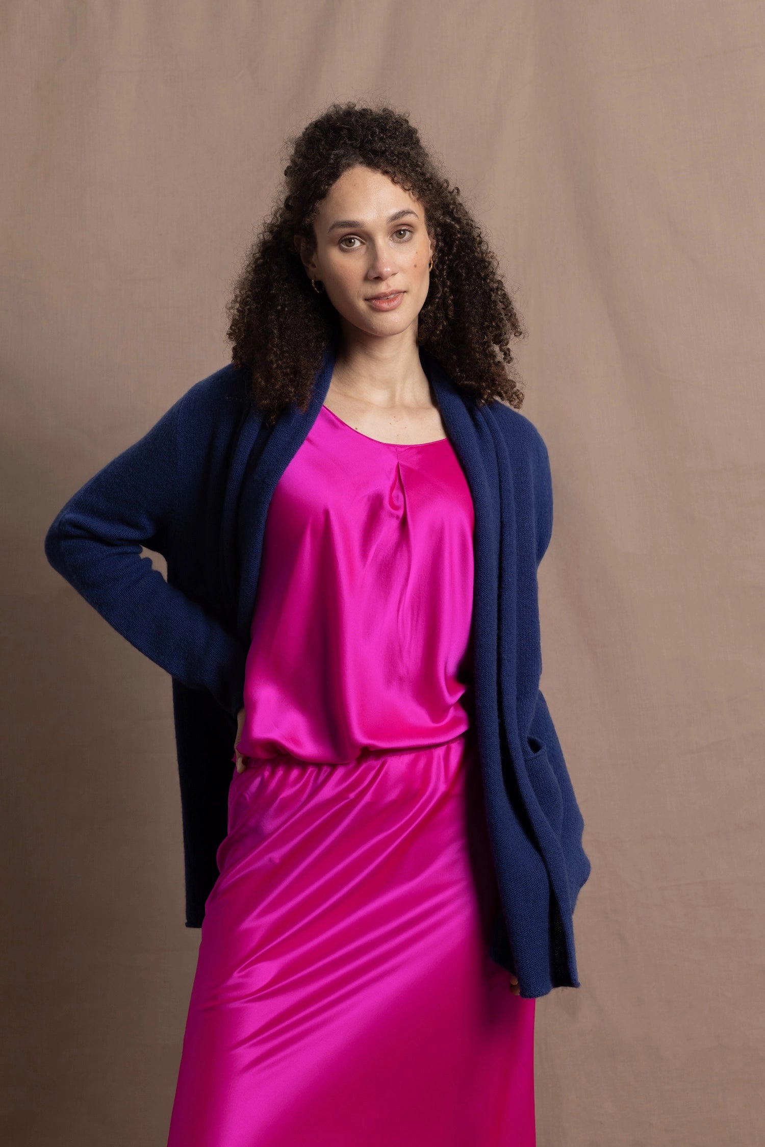 A woman wears an open style longer cashmere cardigan in blue worn over a bright pink 100% silk top and matching skirt