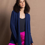 A woman wears an open style longer cashmere cardigan in blue worn over a bright pink 100% silk trousers and navy bodysuit