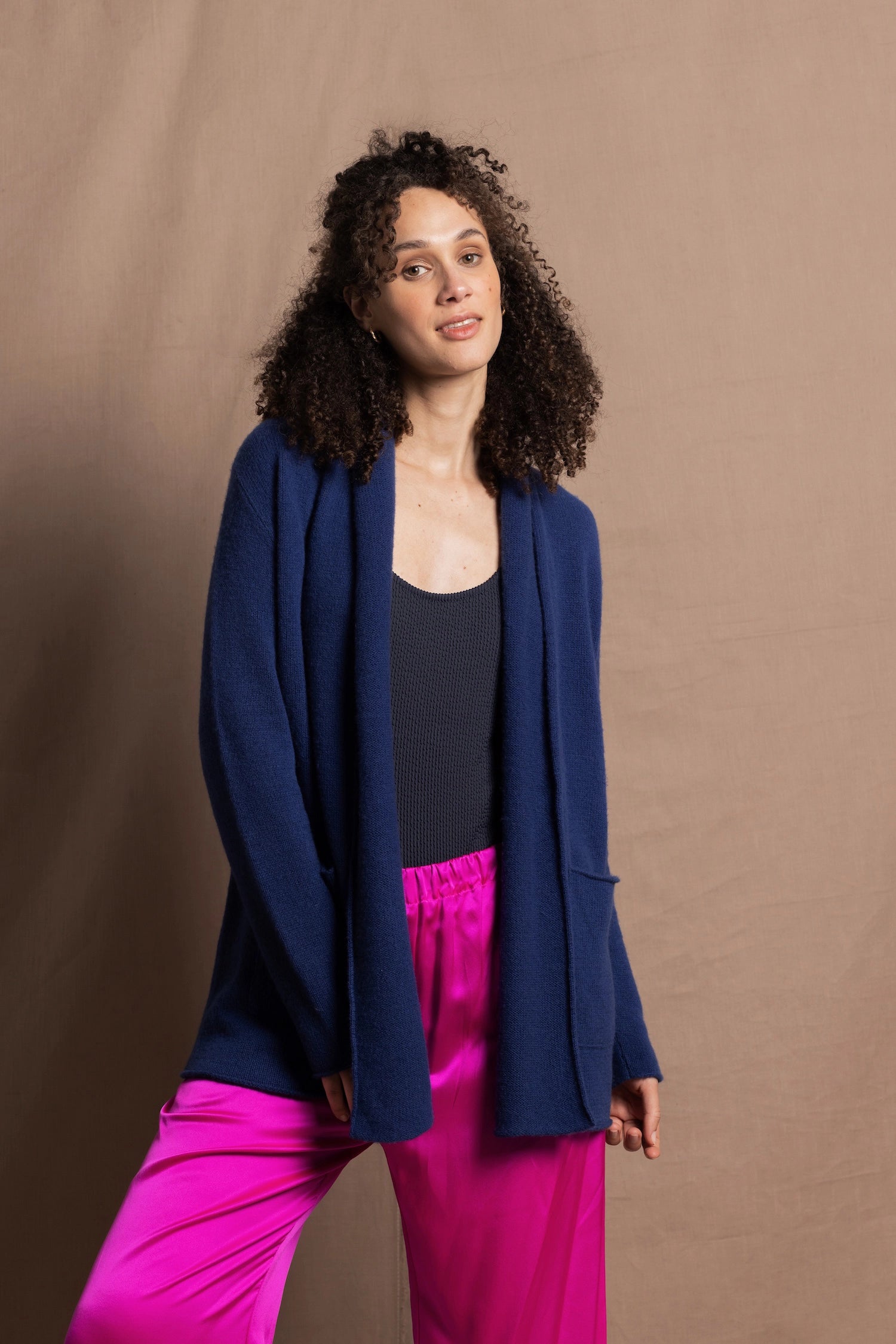 A woman wears an open style longer cashmere cardigan in blue worn over a bright pink 100% silk trousers and navy bodysuit
