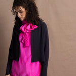 A woman wears a 100% silk pussy bow top in bright pink with matching trousers and a navy cropped cashmere cardigan on top