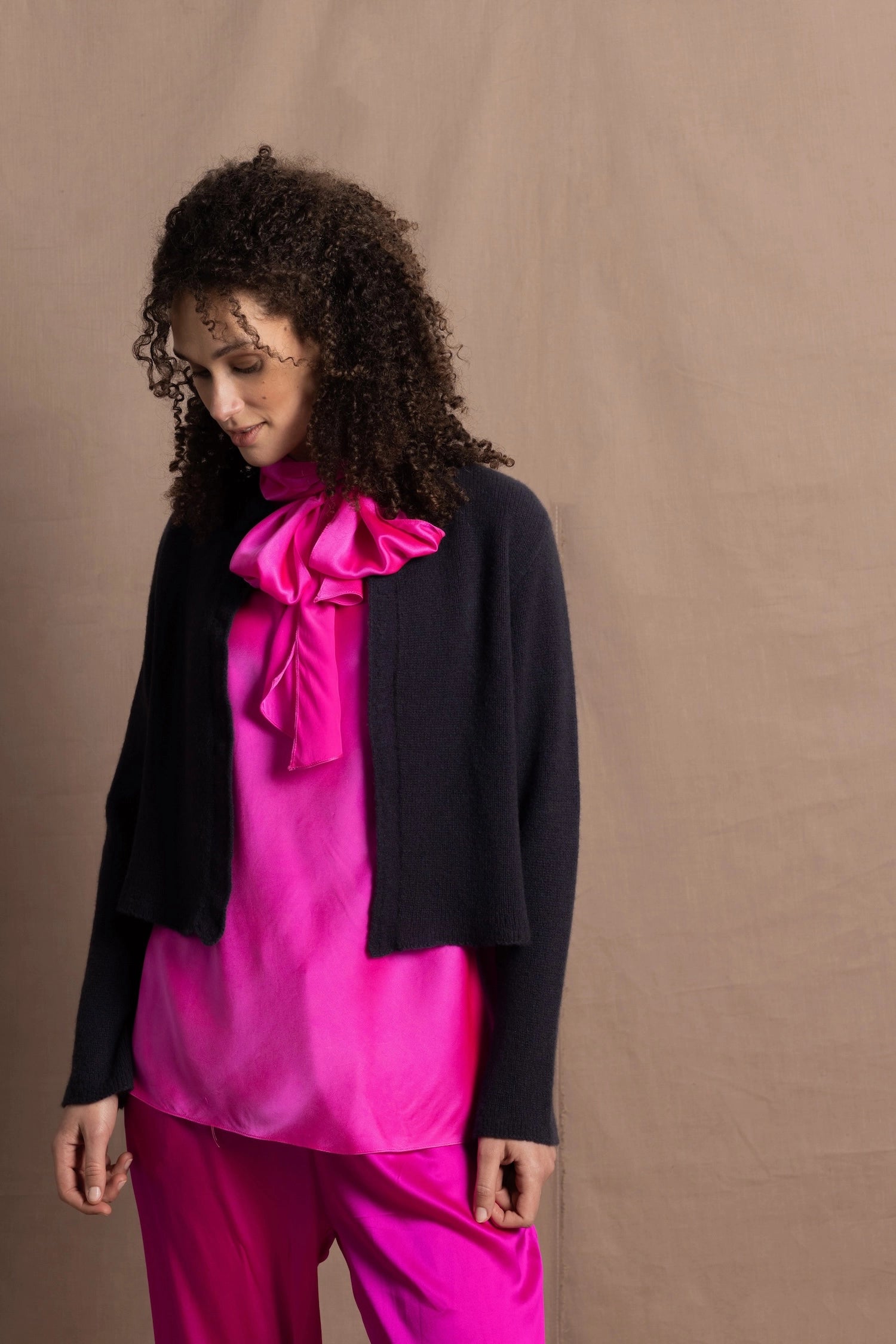 A woman wears a 100% silk pussy bow top in bright pink with matching trousers and a navy cropped cashmere cardigan on top