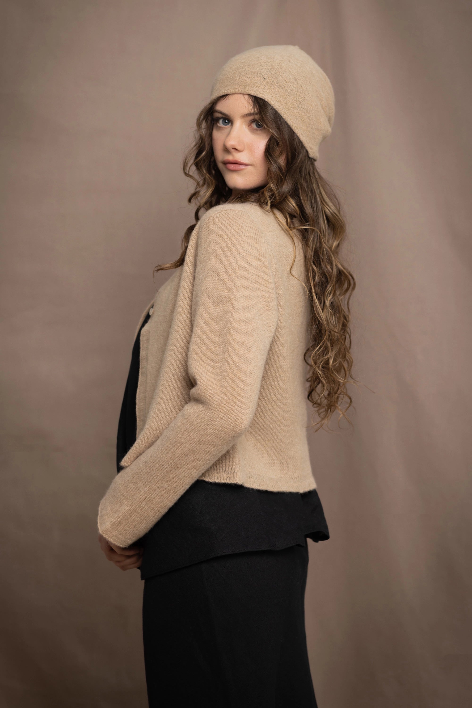 a woman wears a cropped cashmere cardigan in a sandy beige colour with matching beanie hat over black linen top and skirt by Irish brand Madigan