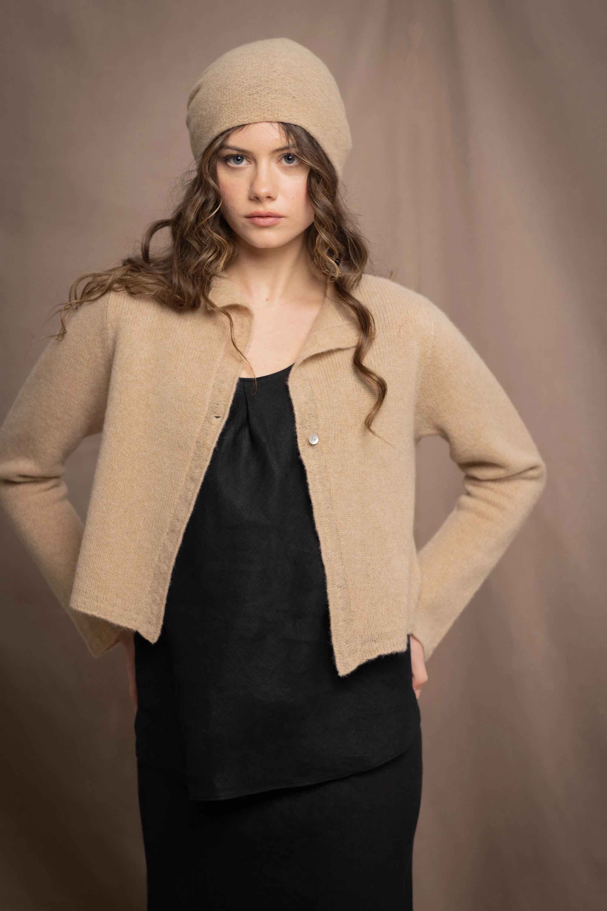 a woman wears a cropped cashmere cardigan in a sandy beige colour with matching beanie hat over black linen top and skirt by Irish brand Madigan