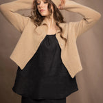 a woman wears a cropped cashmere cardigan in a sandy beige colour with matching beanie hat over black linen top and skirt by Irish brand Madigan