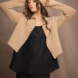 a woman wears a cropped cashmere cardigan in a sandy beige colour with matching beanie hat over black linen top and skirt by Irish brand Madigan