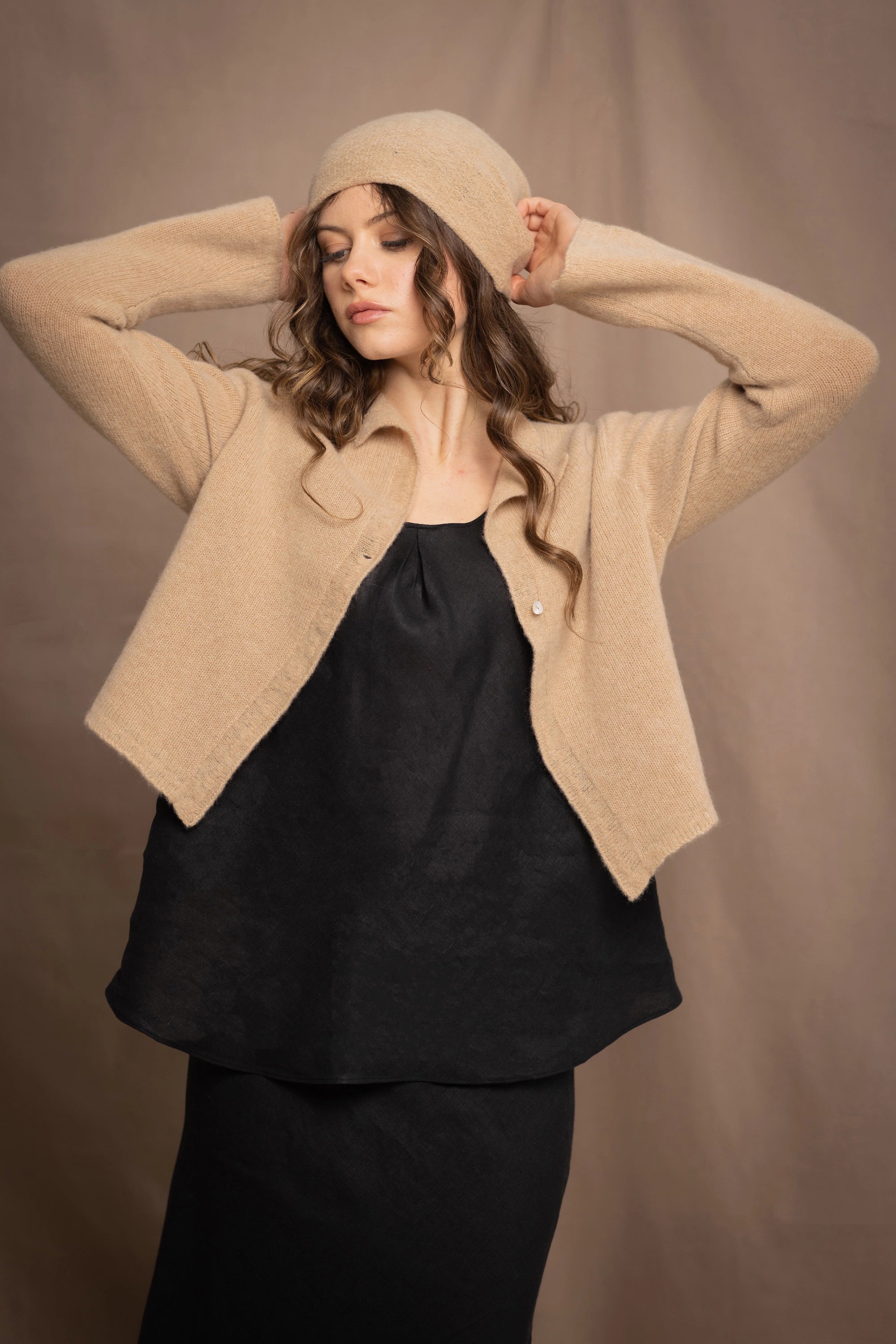 a woman wears a cropped cashmere cardigan in a sandy beige colour with matching beanie hat over black linen top and skirt by Irish brand Madigan