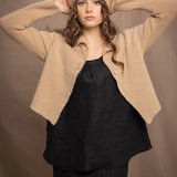 a woman wears a cropped cashmere cardigan in a sandy beige colour with matching beanie hat over black linen top and skirt by Irish brand Madigan