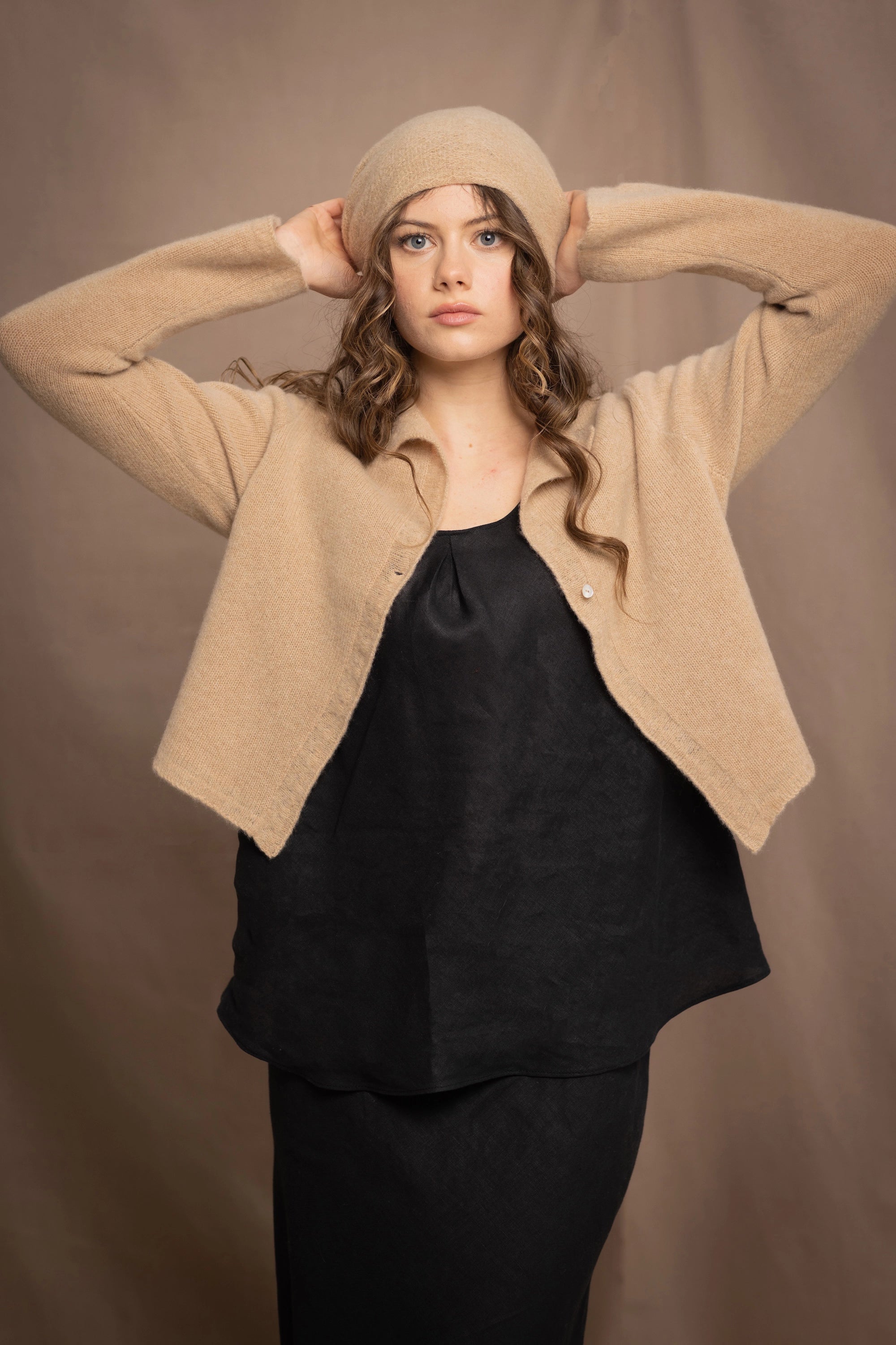 a woman wears a cropped cashmere cardigan in a sandy beige colour with matching beanie hat over black linen top and skirt by Irish brand Madigan