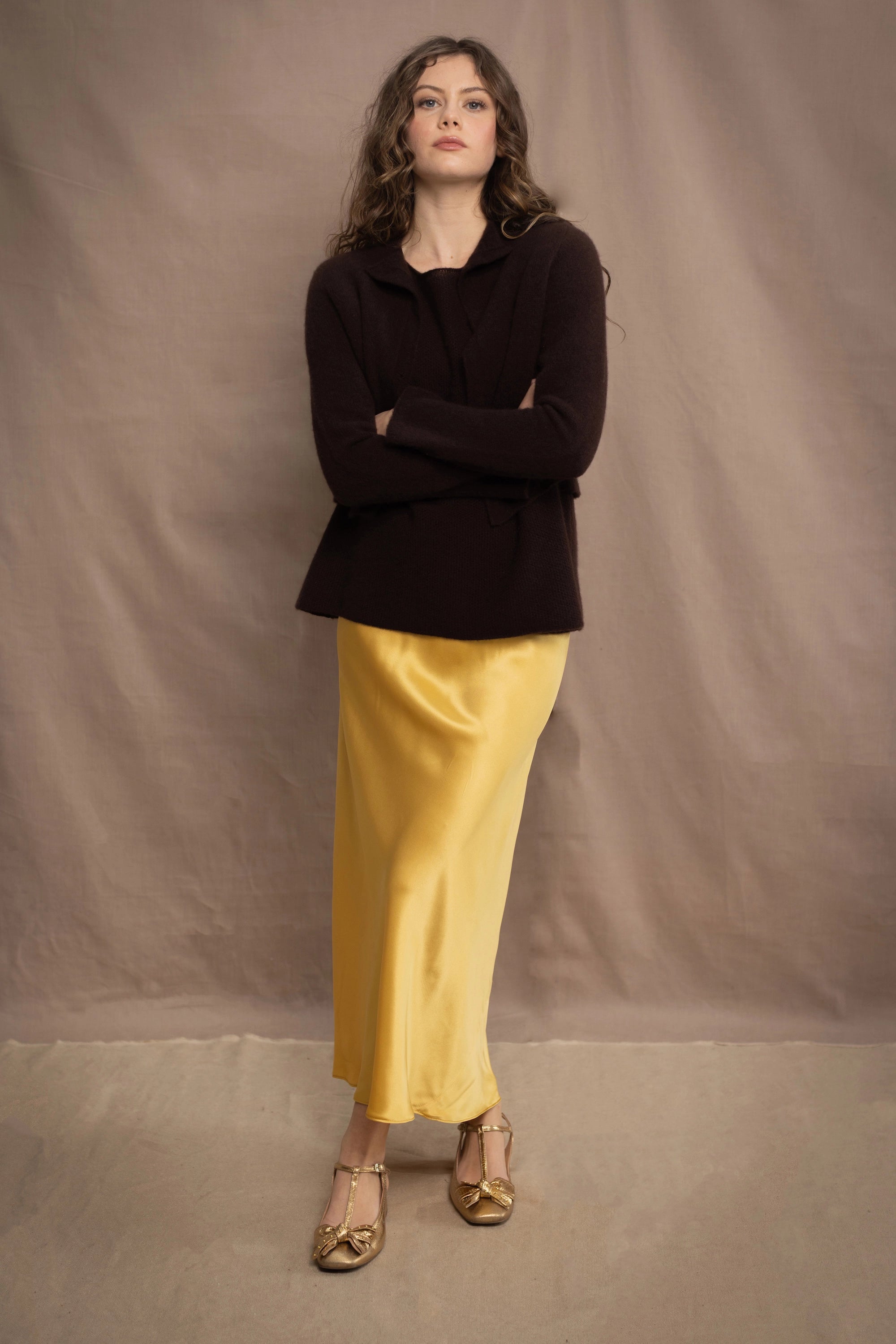 a woman wears a cropped cashmere cardigan in chocolate brown over a matching cashmere tee sweater with a silk skirt in pale yellow