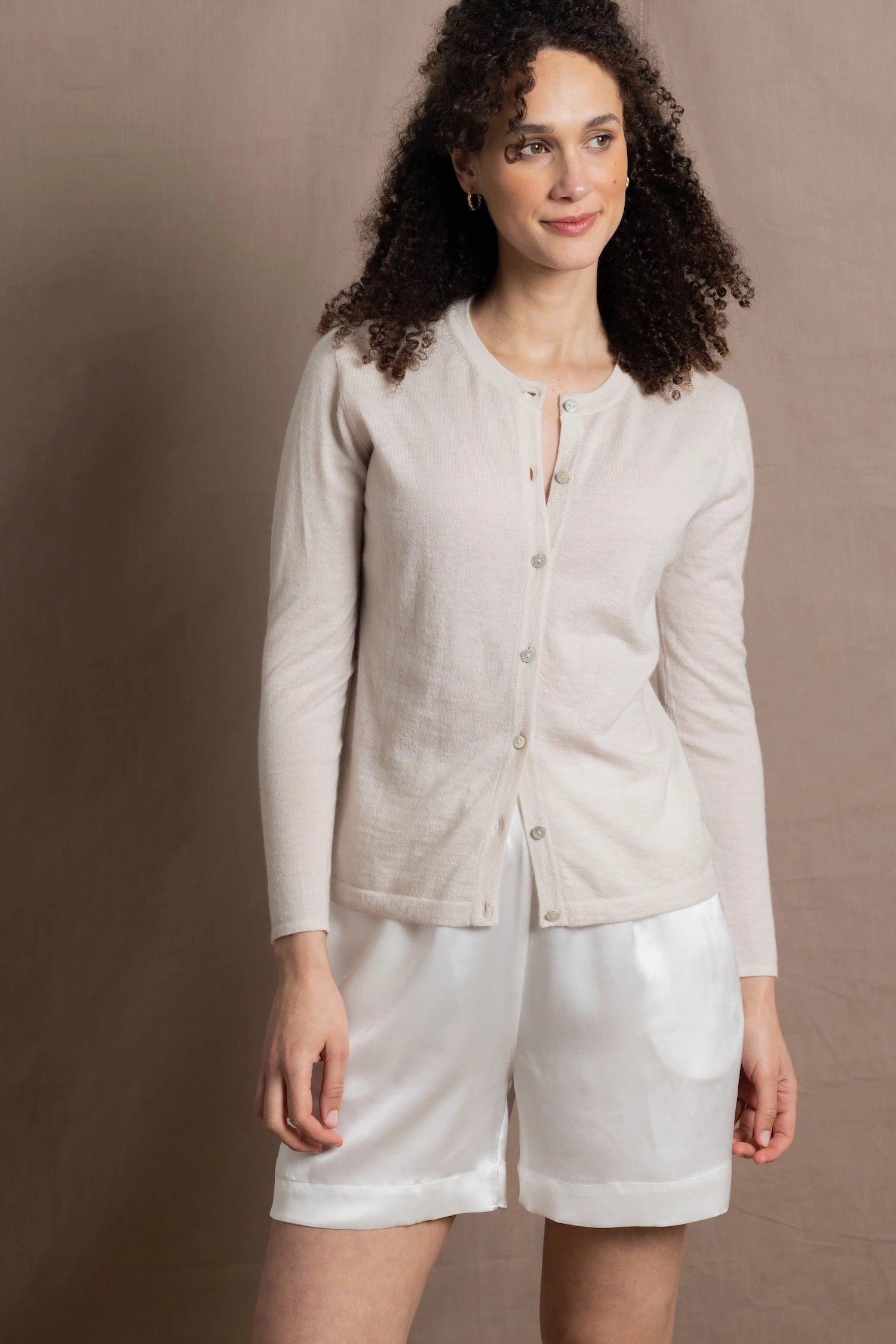 a woman wears a thin summer weight cashmere  button down cardigan sweater in a cream colour with white silk shorts