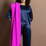 A woman wears a large neon pink cashmere wrap over her shoulder over a full teal silk to and trouser outfit